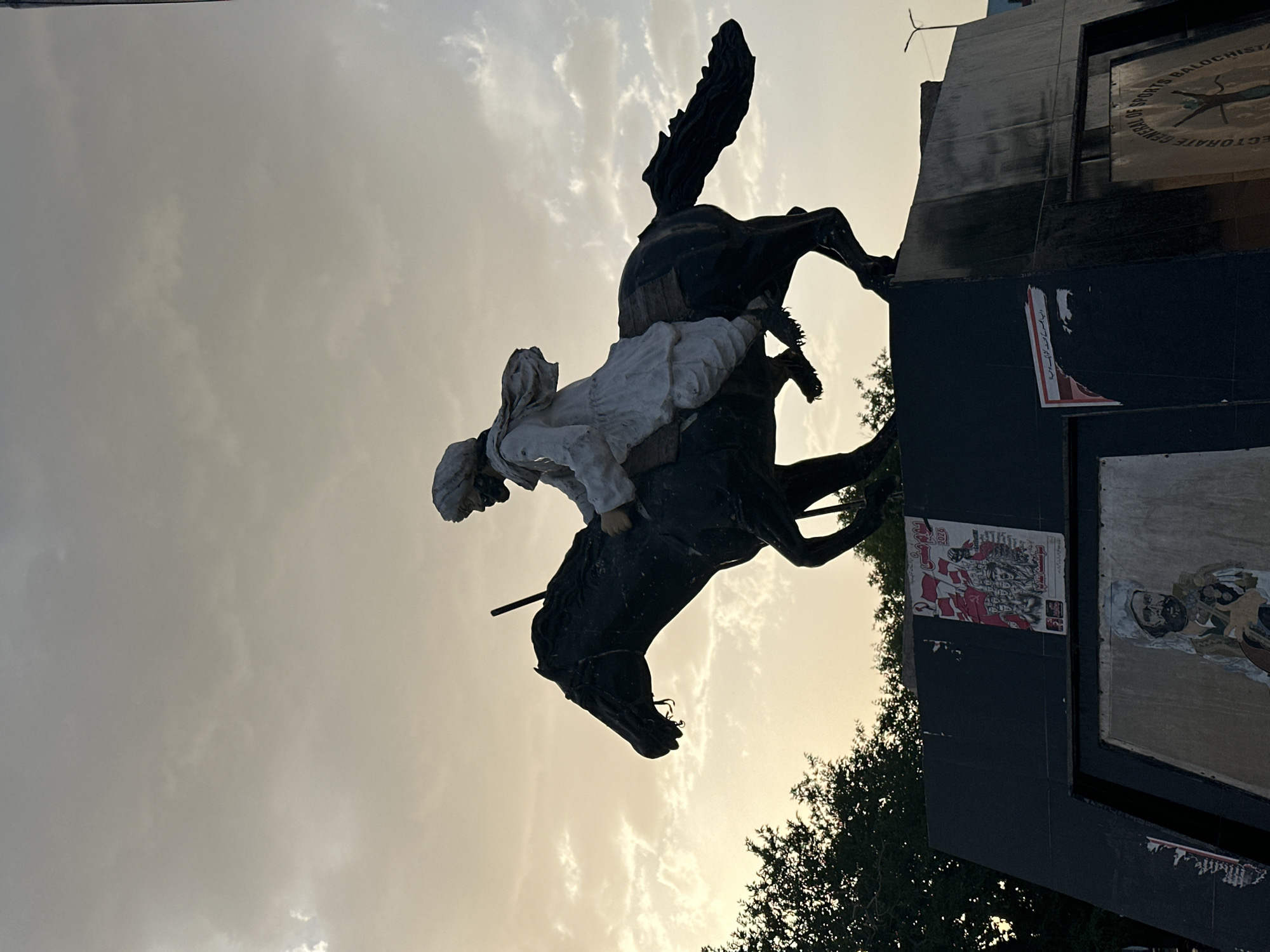 A statue of a person on horseback stands in Quetta City Tehsil, Pakistan. The statue, mounted atop a black, multi-tiered base, depicts a person wearing a white outfit riding a black horse in a rearing position. The base of the statue has various posters and a clock with an emblem attached. Trees are visible at the base of the statue. The background consists of a cloudy sky with a warm, hazy light.