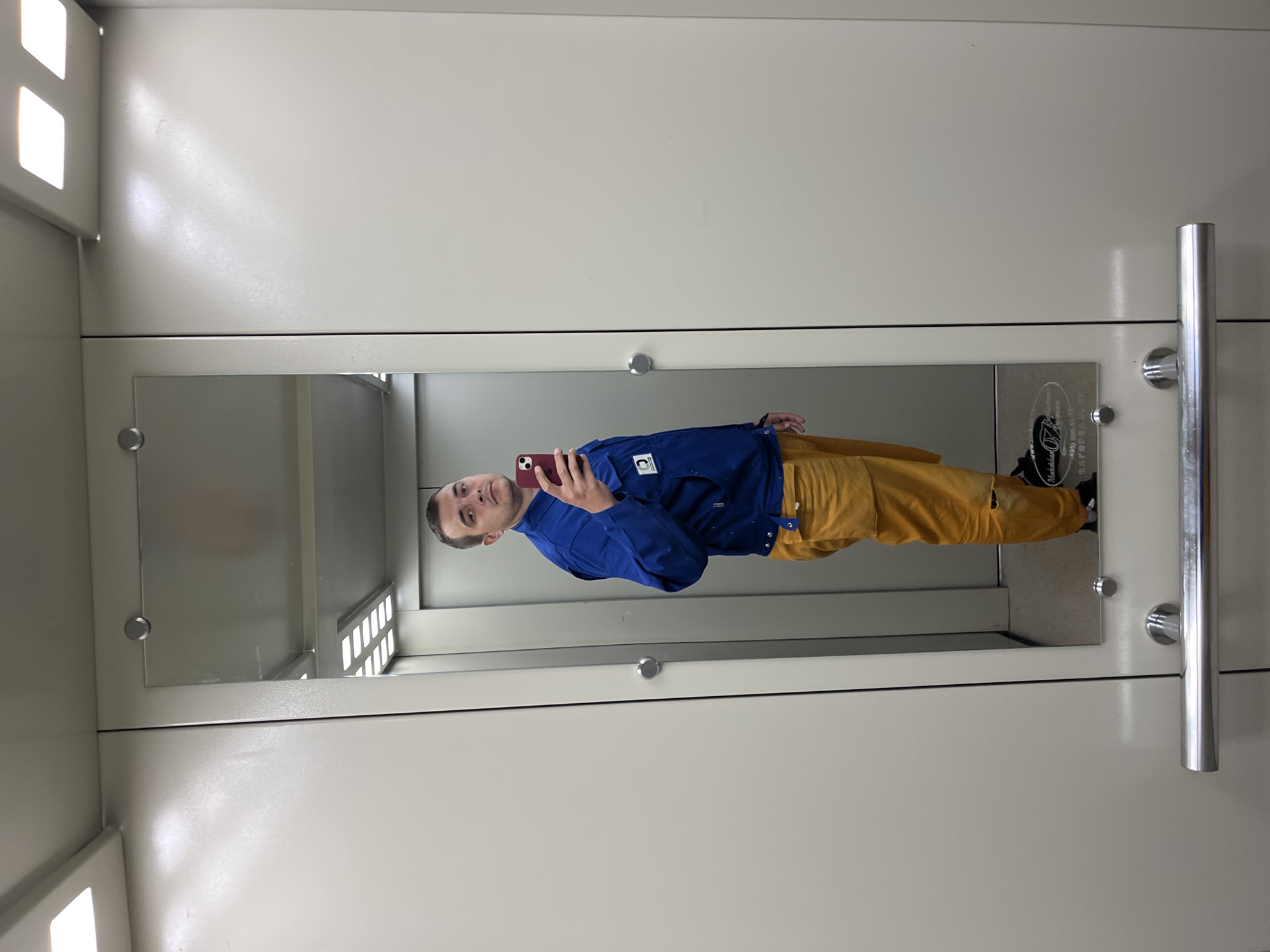 A man is seen in a mirror taking a selfie with a red mobile phone. He is wearing a blue jacket and mustard-colored pants. He appears to be inside a metal changing room with a mirror attached to the door. The location is Mokshino, Russia.