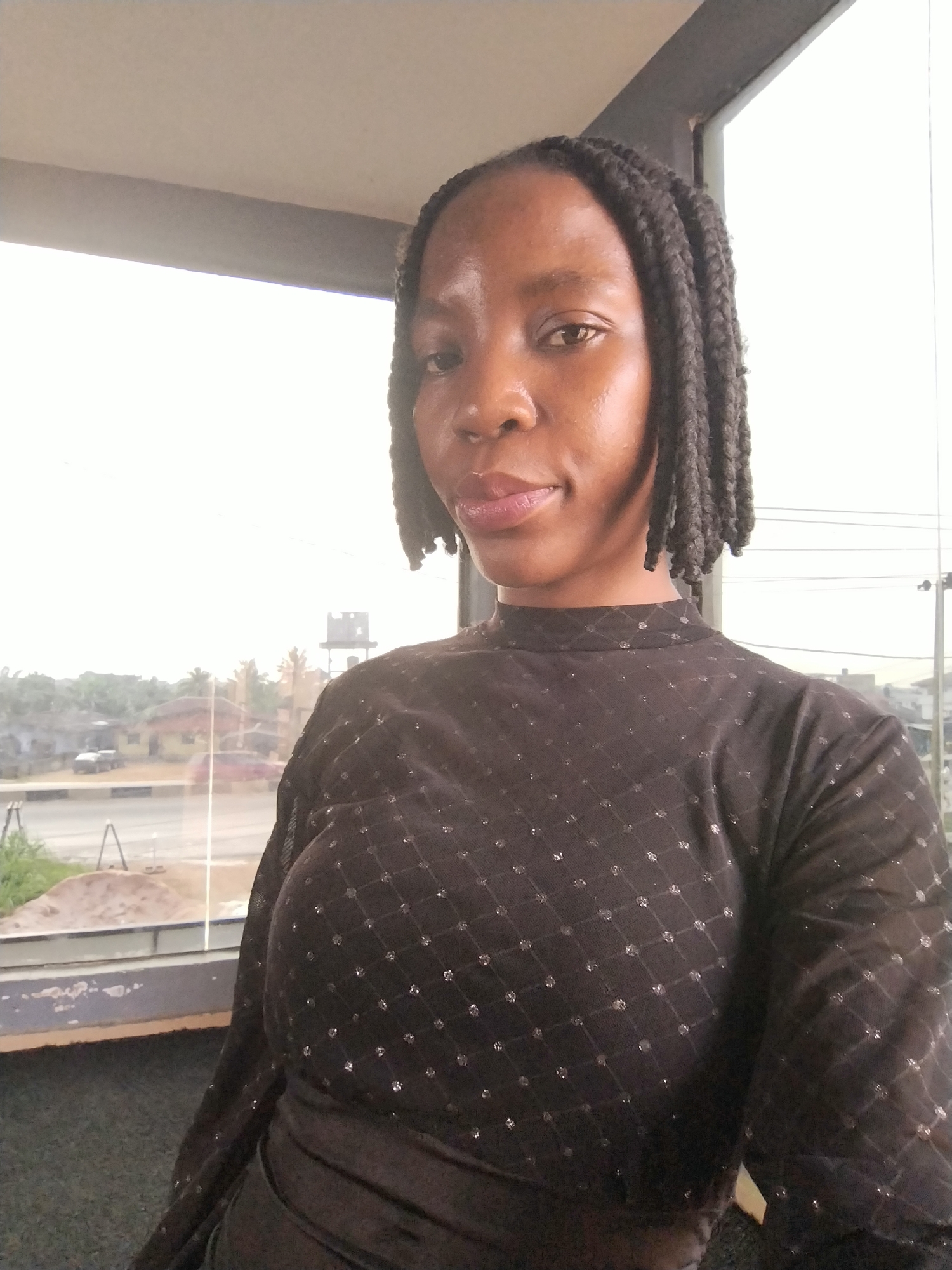 A medium shot shows a Black woman with braided hair from the shoulders up, standing indoors and facing the camera. She wears a dark, long-sleeved shirt with a high neckline, adorned with a subtle diamond-shaped pattern of small, shimmering dots. The background features a large window that looks out onto an overcast urban landscape with buildings, trees, a road, and a water tower. A concrete ledge runs along the bottom of the window. The lighting is soft and diffused, with the window providing the primary light source.