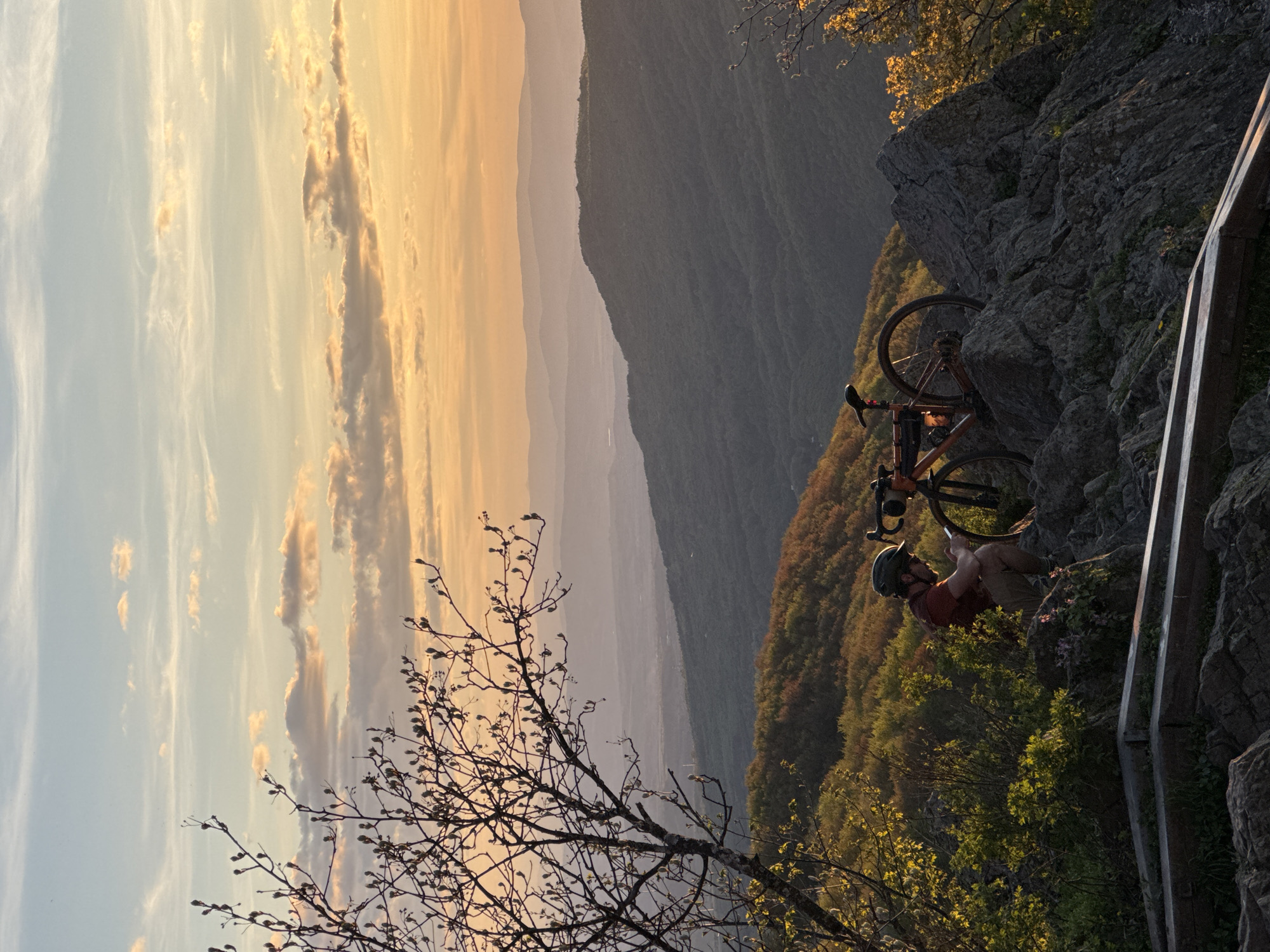 A cyclist rests with his bicycle on a rocky precipice overlooking a vast landscape during a vibrant sunset. The sky transitions from soft blue to a deep golden hue, illuminating distant mountain ranges shrouded in a gentle haze. A leafless tree branch juts into the foreground, its delicate twigs silhouetted against the colorful sky. The cyclist is clad in a red shirt and helmet. The location is not discernible as being specifically within the city of Sofia, Bulgaria.