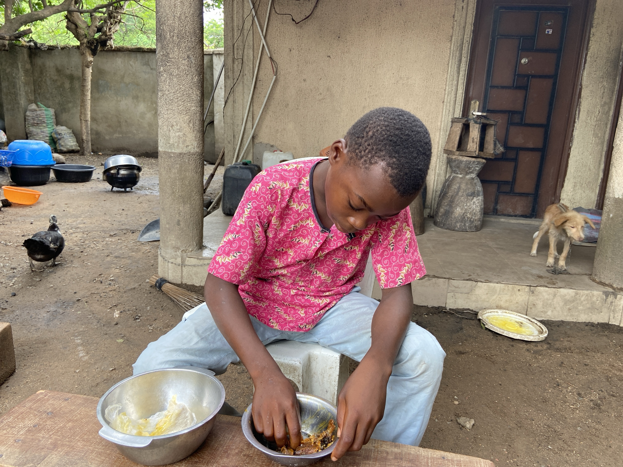 An adolescent male with short, dark hair wearing a pink patterned short-sleeve shirt and light blue jeans is seated outdoors on a small concrete block at Mararaba, Nigeria. He is bent over, his hands inside a small metal bowl containing dark food matter. To his left is a larger metal bowl filled with light-colored contents. Both bowls rest on a wooden surface.
Behind him, a beige concrete building with a brown door and decorative metal bars is visible. A wooden structure rests on a stone base beside the door. A tan dog stands on the concrete step in front of the door. To the left of the boy, a black duck is on the dirt ground. In the background, blue and black tubs, a cooking pot, bags of materials, and a metal ladder can be seen. A broom rests against a concrete pillar. The ground is dirt, and a yellow-tinged bowl lies on the ground to the right. Overhead, power lines and a tree are visible.