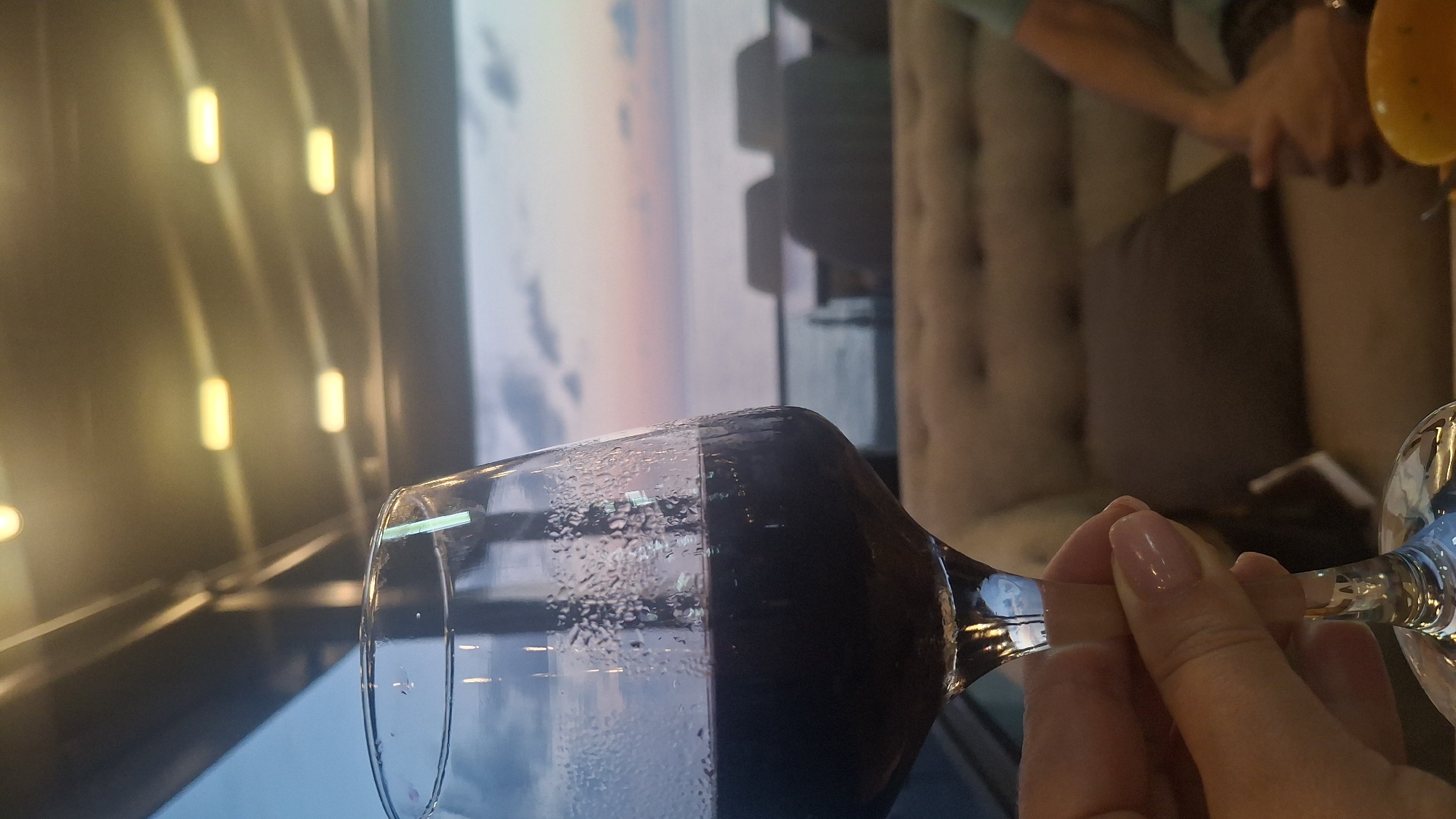 A close-up, eye-level shot shows a hand holding a stemmed wine glass filled with red wine. The glass is partially obscured by condensation. In the background, an outdoor seating area is visible, with plush seating and tables. Beyond the seating area, a body of water, likely the Black Sea, stretches to the horizon under a twilight sky with soft pink and blue hues. The lights of an overhead structure are visible, casting a warm glow. This scene is set at the Black Sea coast in Kobuleti, Georgia.