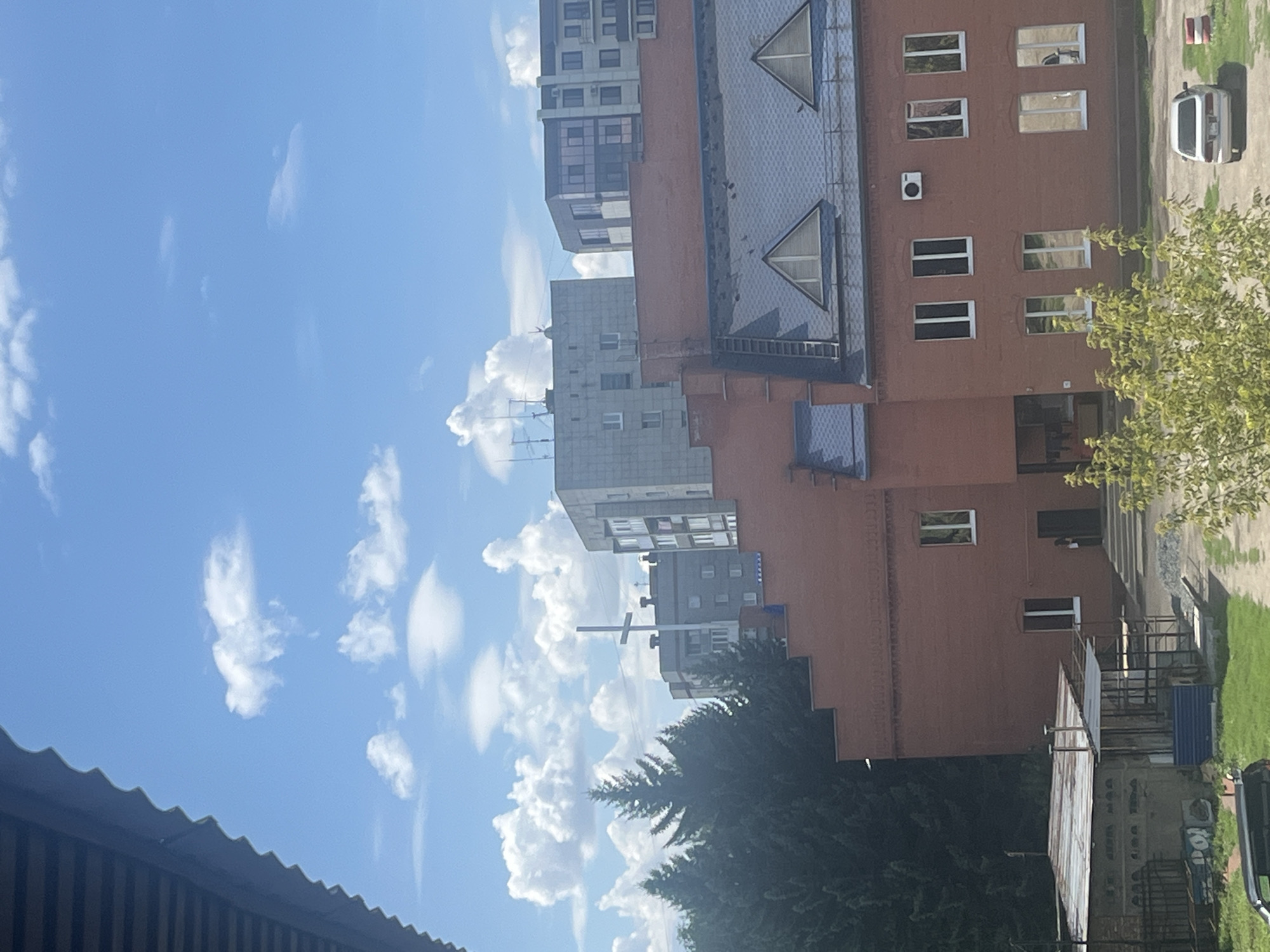 The image captures a view from a building in the city of Oskemen, Kazakhstan. The foreground is partially obscured by the corrugated metal roof of an overhang. A large, red brick building with multiple windows and a dark, sloped roof dominates the mid-ground. Several of these windows are large and rectangular, some reflecting the sky. On the roof of this building, there are at least two triangular dormer windows and a ladder leading up the side. A white air conditioning unit is mounted on the exterior wall. A smaller, dilapidated building with a corrugated metal roof and a fenced area is visible to the left.
In the background, evergreen trees are densely packed on the left side of the frame. Behind the trees and buildings, several multi-story apartment buildings are visible, one with a prominent white cross on its roof. The sky is a vibrant blue, dotted with fluffy white cumulus clouds. A light-colored sedan is parked in a paved area in the lower right corner of the image, next to a white and red traffic barrier. The scene appears to be an outdoor urban environment on a clear, sunny day.