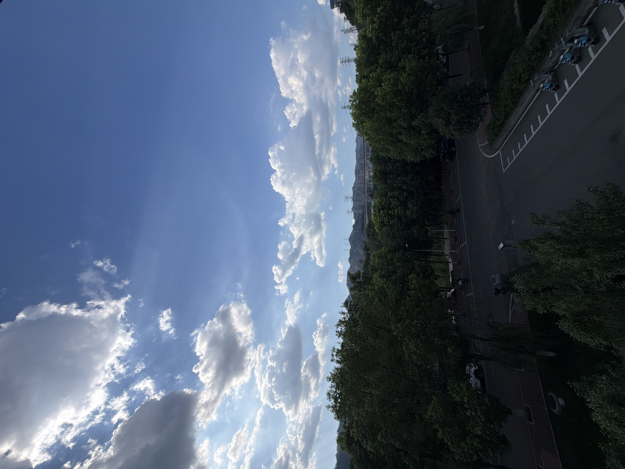 The image depicts a wide-angle view of a suburban or park-like area, likely in Jiangjin, China, characterized by lush green trees and a clear blue sky dotted with fluffy white clouds. The foreground is dominated by dense trees, with a road winding through the scene. Several parked electric scooters are visible along the side of the road, indicating a modern urban or semi-urban setting. In the background, power lines and distant hills suggest the presence of infrastructure and natural landscapes. The lighting indicates it is daytime, possibly late afternoon, with sunlight streaming through the clouds, creating a somewhat dramatic sky. There are no people clearly visible in the image, but the presence of parked vehicles and the road suggests human activity.