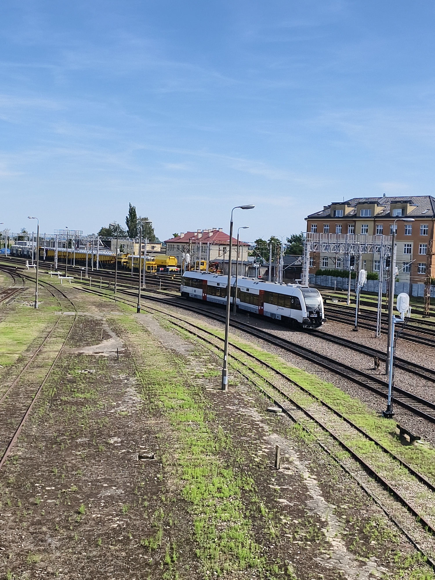 The image shows a train station in Tczew, Poland, on a sunny day. A modern, white and red passenger train is on the tracks in the foreground, traveling from right to left. In the background, there are several yellow freight trains parked on parallel tracks. The station has multiple sets of railway tracks, overhead power lines, and signal poles. Buildings with red roofs are visible behind the tracks. The ground surrounding the tracks is a mix of gravel and grass. The sky is clear and blue.