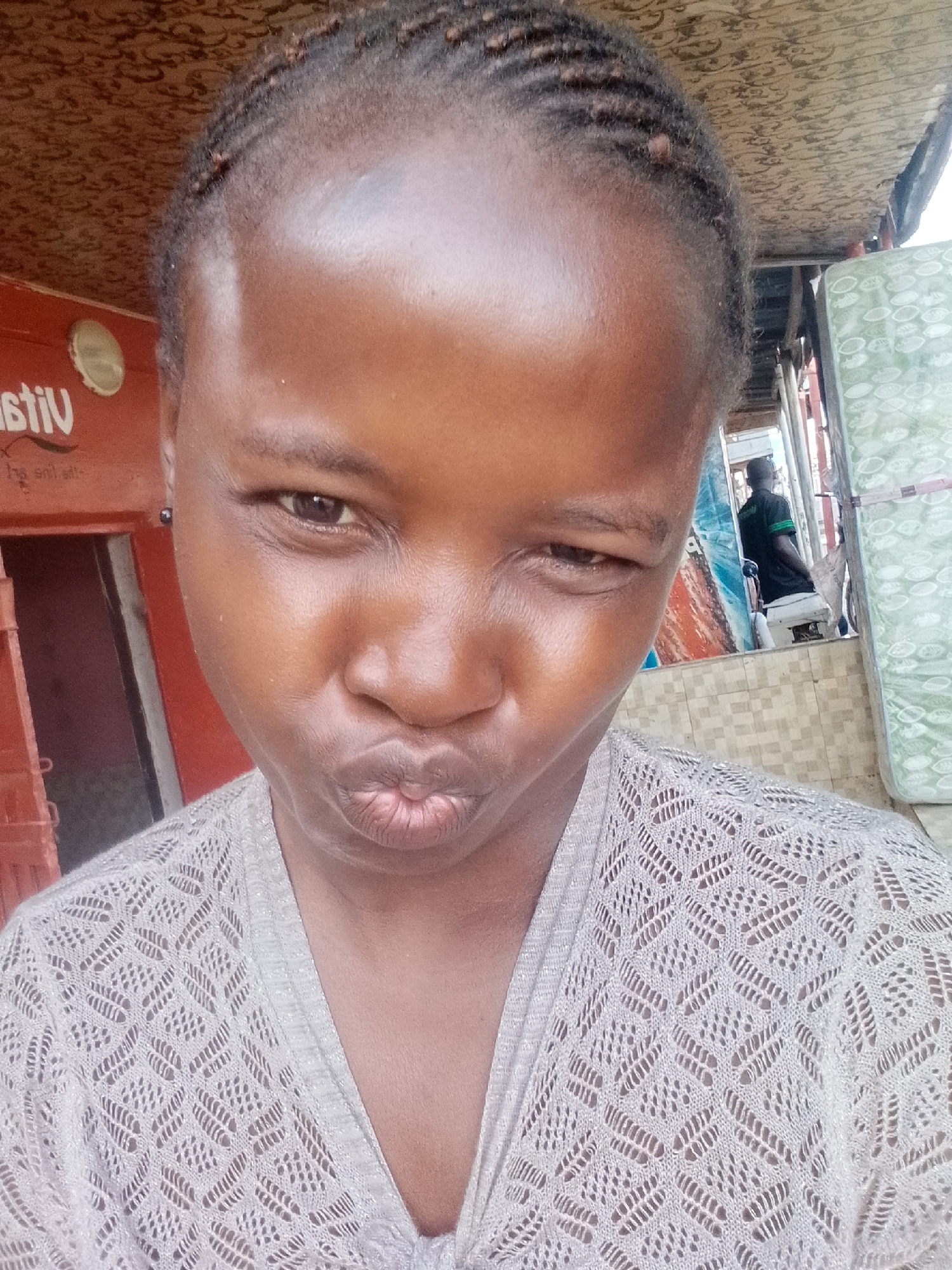 A young Black female with braided hair and a light-colored, patterned sweater is the subject of this close-up, eye-level, outdoor selfie. She is positioned in the center of the frame, with her head tilted slightly and her lips pursed. Her right eye is closed in a wink. The background suggests a street scene with a red wall to the left, featuring a white circular logo with the word "vit" visible. To the right, a patterned mat is draped over a structure, and a man in a dark shirt is visible in the mid-ground. The lighting is natural, indicating it is daytime. The location is identified as Pantame, Nigeria.