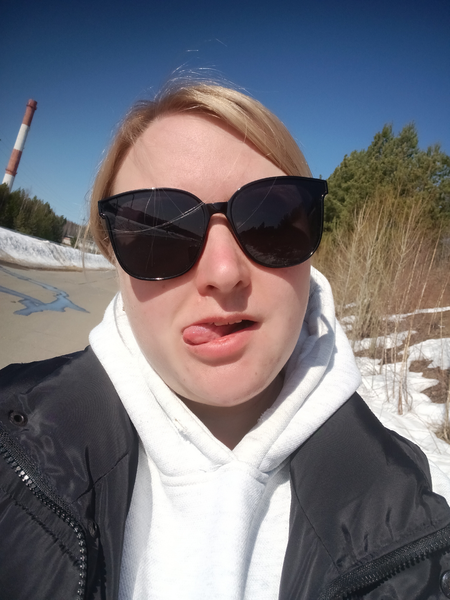 A fair-skinned person with blonde hair, wearing black sunglasses with a visible crack in the left lens, a white hooded sweatshirt, and a black jacket, is sticking their tongue out at the camera. The photo appears to be a selfie. In the background, there's a paved road with some snow on the sides, trees, and a tall industrial chimney with red and white stripes. Based on the geographical cues, the location is likely Chaikovsky, Russia. The overall scene appears to be outdoors on a sunny day in a location where winter is receding, given the presence of snow.