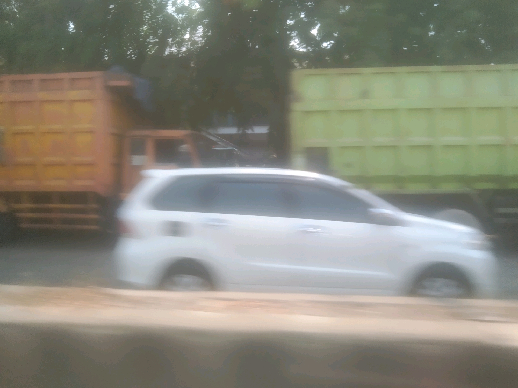The image depicts a blurry street scene in Bandar Lampung, Indonesia, likely taken from a moving vehicle. In the foreground, a beige, elevated barrier is visible. Behind it, a white minivan is driving, its wheels and sides blurred by motion. To the left of the minivan, a large, orange truck with a flatbed is parked or moving slowly. Further to the right, a light green truck with a similar flatbed is also visible. Trees and foliage line the background, indicating an outdoor setting. The overall impression is of traffic on a street in a tropical or subtropical environment.