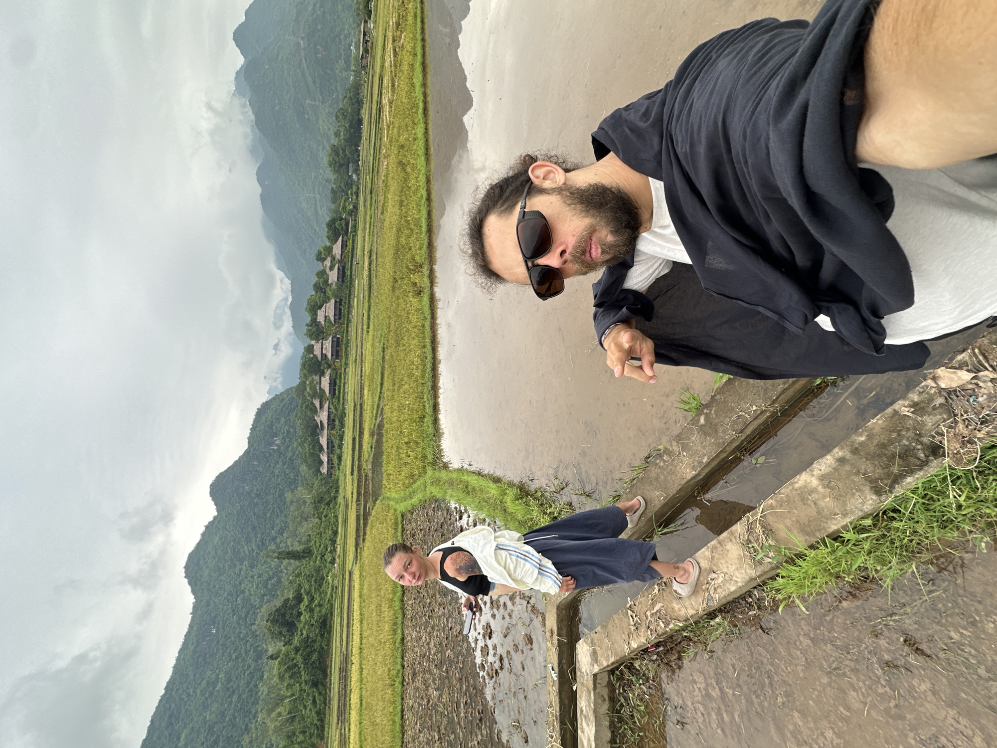Two individuals are captured in a selfie taken in the rice paddies of Mai Chau, Vietnam. The man in the foreground, positioned on the right, wears sunglasses and a black wrap over a white shirt, with his left arm extended towards the camera. To the left, a woman stands on a concrete pathway, looking back at the camera. She has dark hair pulled back and is wearing a black top, a white jacket, and dark blue pants. Behind them, the landscape unfolds with vibrant green and yellow rice fields, reflecting the overcast sky. In the distance, lush, mist-shrouded mountains rise, dotted with traditional stilt houses. The ground appears wet and muddy, indicative of the agricultural setting.