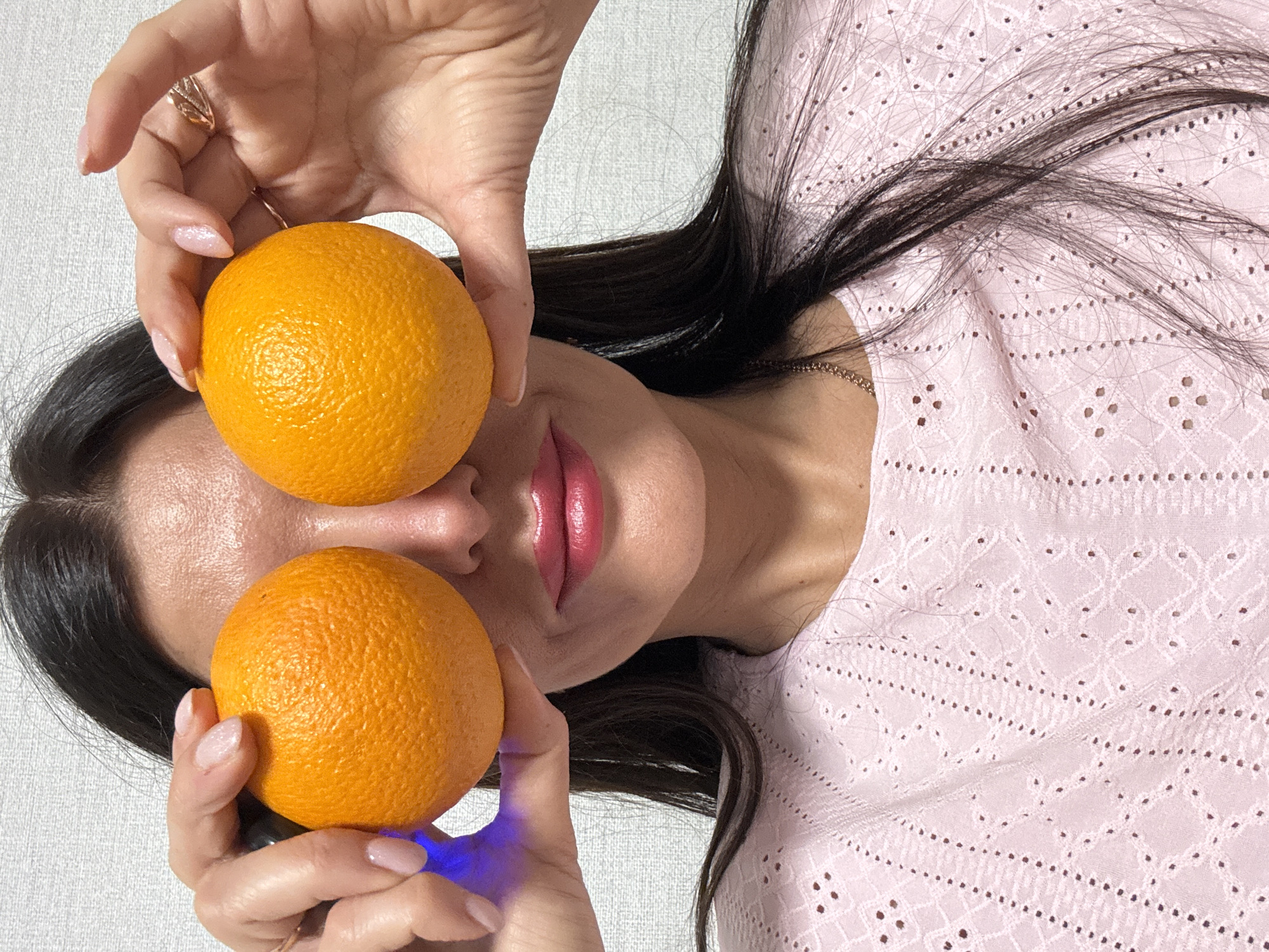 A woman with long dark hair is holding two oranges in front of her eyes. She is wearing a pink top with a patterned design. The background is a light gray or white wall. The image appears to be taken indoors. There is no clear evidence to suggest this image was taken in Chaikovsky, Russia.