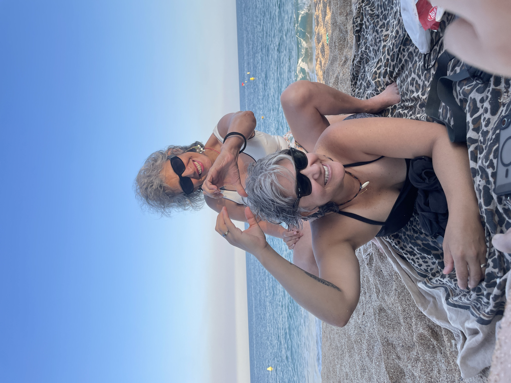 Two women with grey hair and sunglasses are posing for a photo on a beach in Canet de Mar, Spain. The woman in the foreground is lying down on a leopard-print blanket, wearing a black bikini top. The woman behind her is kneeling, wearing a white tank top. They are both making heart shapes with their hands. The ocean is visible in the background, with a clear blue sky above. The beach is sandy, with some small waves breaking near the shore. There are yellow buoys in the water.