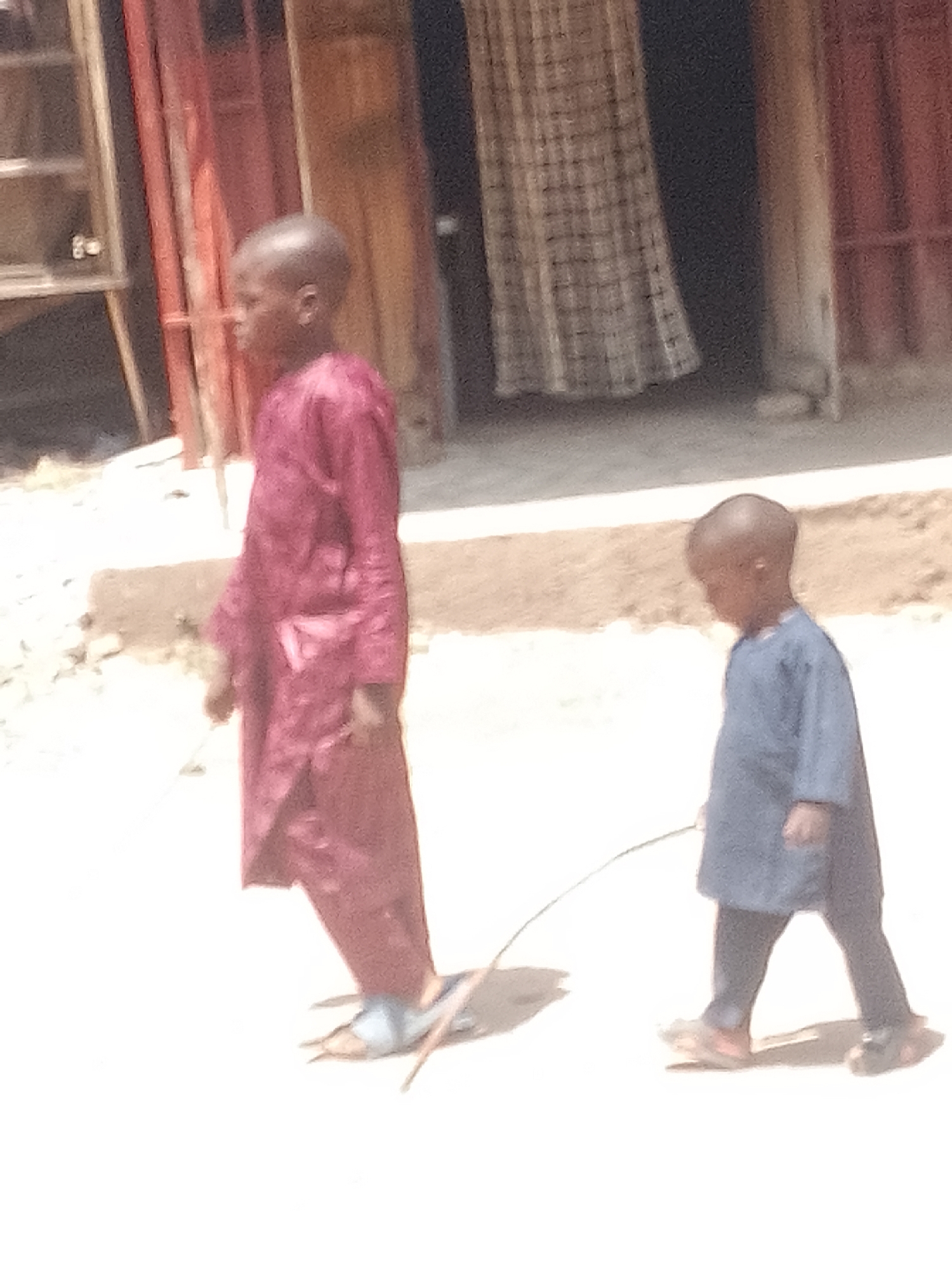 Two young boys walk on a dusty, sunlit outdoor path in Pantame, Nigeria. The boy on the left, wearing a long, dark red robe and sandals, walks away from the camera. He is holding a thin stick. The boy on the right, wearing a blue tunic and dark pants, walks beside him, also holding a stick. They appear to be moving in the same direction. Behind them, a building with red and brown facades and a checkered curtain is visible. The overall lighting suggests a bright, daytime scene.