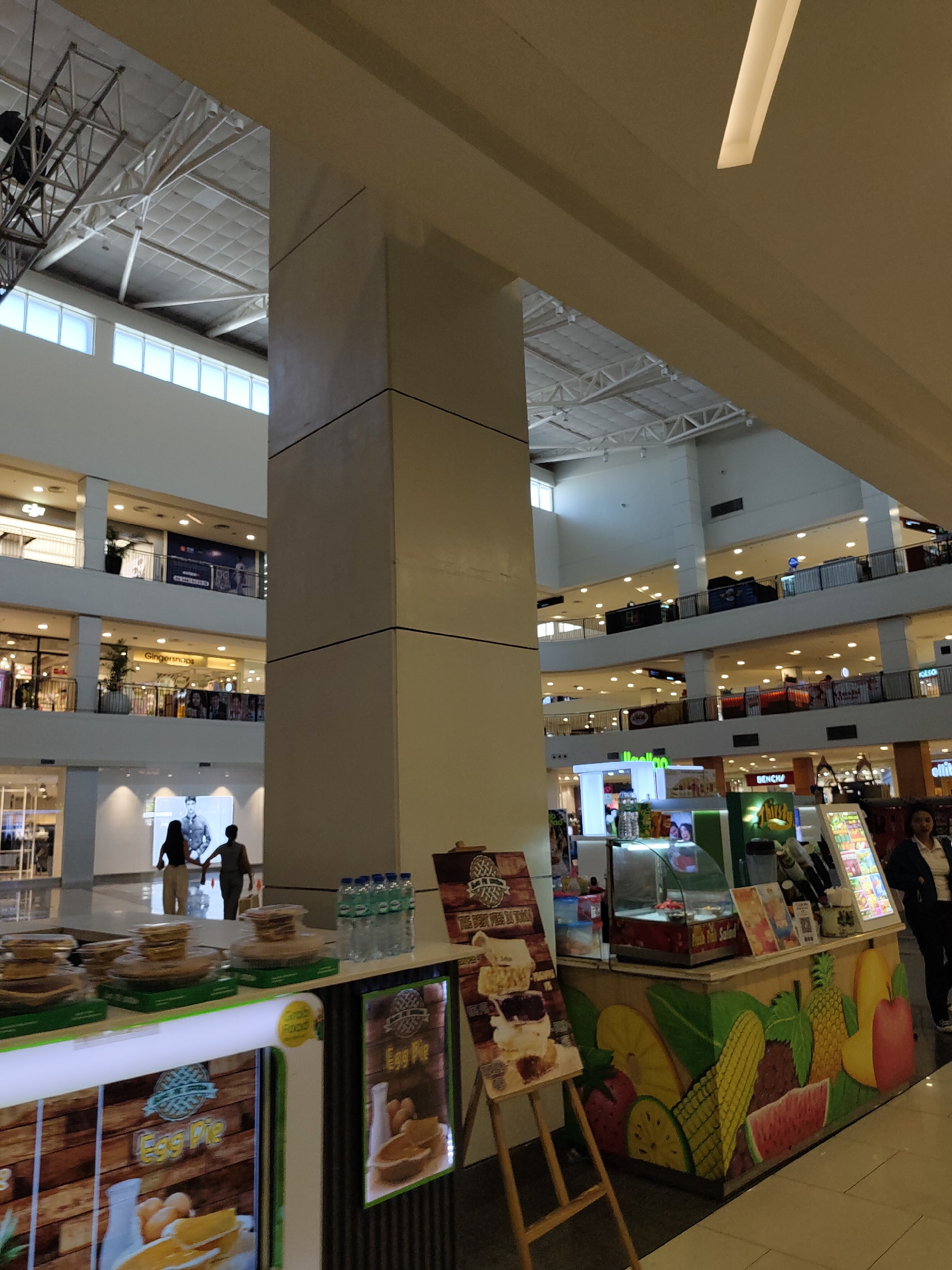 The image shows a bright, multi-level indoor shopping mall, likely in Davao City, Philippines, with various shops visible on different floors. In the foreground, a food kiosk is set up, featuring colorful graphics of fruits like pineapples, watermelons, and apples. The kiosk displays an assortment of food items, including what appear to be pastries or pies, and bottled water. To the left of the kiosk, a stand displays a poster for "Egg Pie." In the mid-ground, two individuals, a man and a woman, are walking away from the camera, hand in hand. The mall's architecture includes large support pillars, a high ceiling with exposed structural elements and lighting, and upper levels with railings and storefronts.