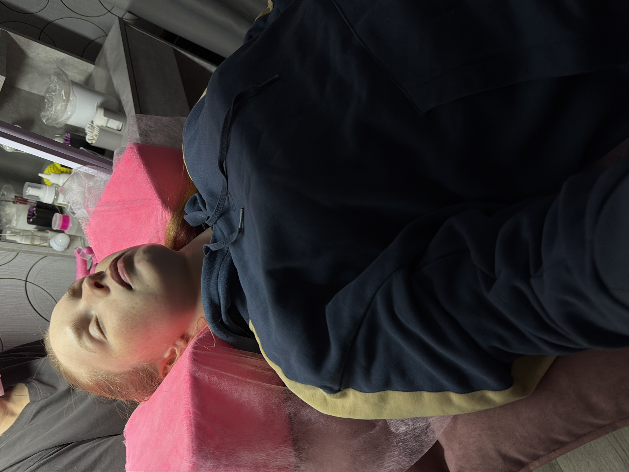 A young person is lying on their back, resting on a pink pillow. They are wearing a dark blue hooded sweatshirt with light yellow trim around the bottom. The person's eyes are closed and their face is visible, revealing light skin and freckles. To the left of the person is a gray or black garment. In the background, there is a shelf holding various cosmetic or personal care products and an obscured gray wall. The scene appears to be indoors, potentially in a salon or spa, but the specific location in the city of Berezniki, Russia cannot be determined based on the image alone.