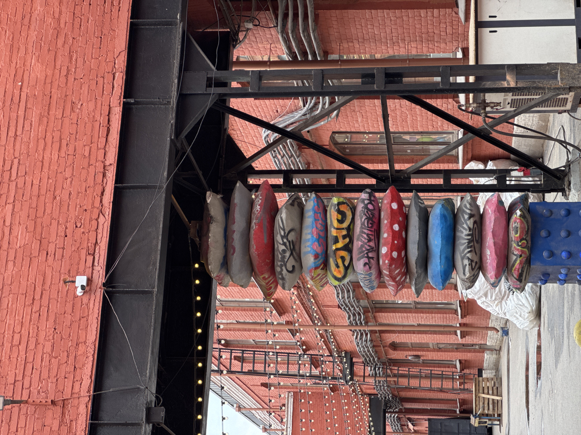 The image shows a street scene likely located in an urban area, characterized by a brick building and industrial infrastructure. A row of painted, boat-shaped objects hangs from a metal structure, potentially a part of an artistic installation or street decoration. The objects are diverse in color and design, with patterns ranging from solid colors to graffiti-like motifs and polka dots.
The architecture includes a prominent brick wall with visible seams and texture. Metal pipes and wiring are exposed along the upper portion of the wall, contributing to the industrial aesthetic. A security camera is affixed to the brick wall, suggesting monitoring of the area.
In the background, there's a building with multiple windows, implying the presence of either residential or commercial spaces. The scene is captured at an angle that emphasizes the verticality of the buildings and the interplay of colors and textures between the brick, metal, and painted objects.
The location is possibly in Saint Petersburg, Russia, due to the architectural style and urban layout that resemble typical Russian cityscapes. However, there isn't specific evidence within the image to definitively confirm this.