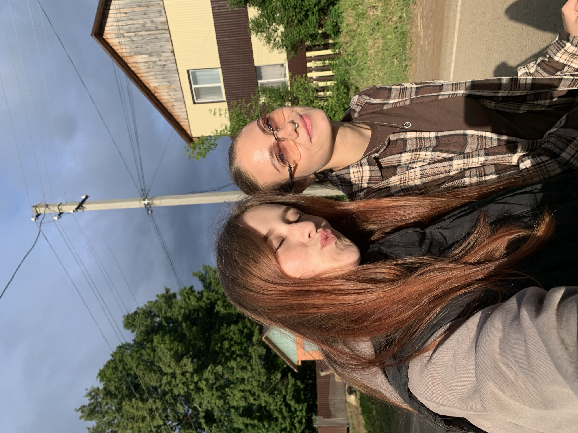This image captures a close-up, tilted outdoor selfie of two young women in Фоки, Russia. The woman in the foreground has long, reddish-brown hair, with her eyes closed and lips playfully pursed in a kissing expression. She is wearing a dark top, possibly a jacket, over a lighter grey long-sleeved garment.
Behind her, and slightly to the left, is another woman with lighter hair, wearing stylish light pink-tinted sunglasses and a nose piercing. She sports a brown plaid button-up shirt over a plain brown t-shirt, accessorized with a silver chain necklace featuring a small circular pendant. Her eyes are open, looking towards the camera with a subtle, calm smile.
The background reveals a rural or suburban setting under a partly cloudy sky. To the left, a prominent utility pole with multiple power lines extends upwards. A light yellow house with a dark brown roof and trim, featuring at least two windows, is visible behind the women. Lush green trees and foliage are scattered throughout the background, and a paved path or road runs along the right side of the frame. The warm lighting suggests it is daytime, possibly late afternoon, with sunlight highlighting the subjects' hair and faces. The overall atmosphere is relaxed and friendly. No visible text is present in the image.