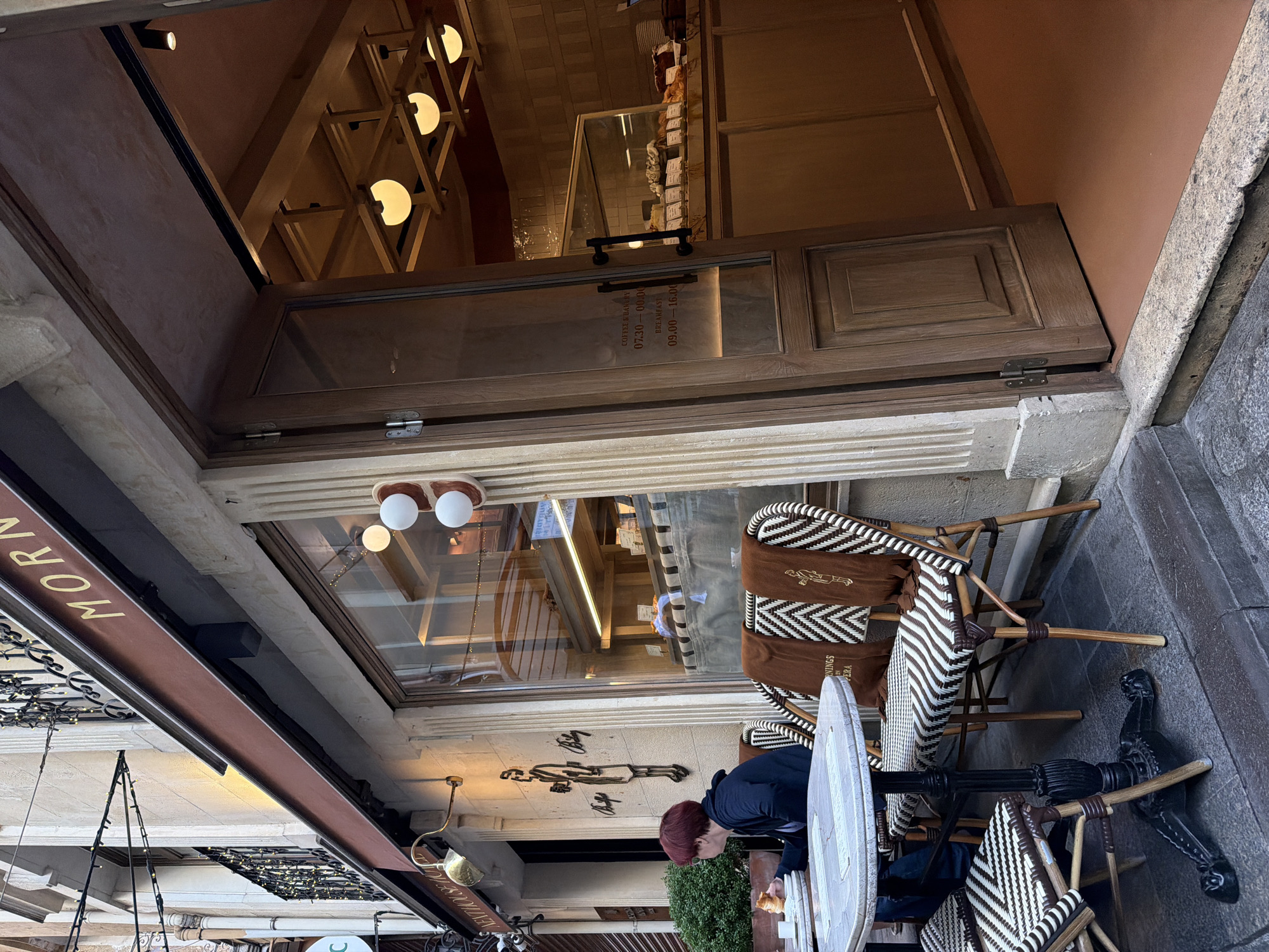 The scene is a view of a cafe exterior in the Beyoğlu district of Istanbul, Turkey. The cafe is partially visible through an open wooden-framed door, with a sign above the door reading "MORNINGS". Tables and chairs, some occupied by patrons, are arranged outside. The cafe appears to have a window display and interior lighting. The overall environment suggests a European-style cafe setting.