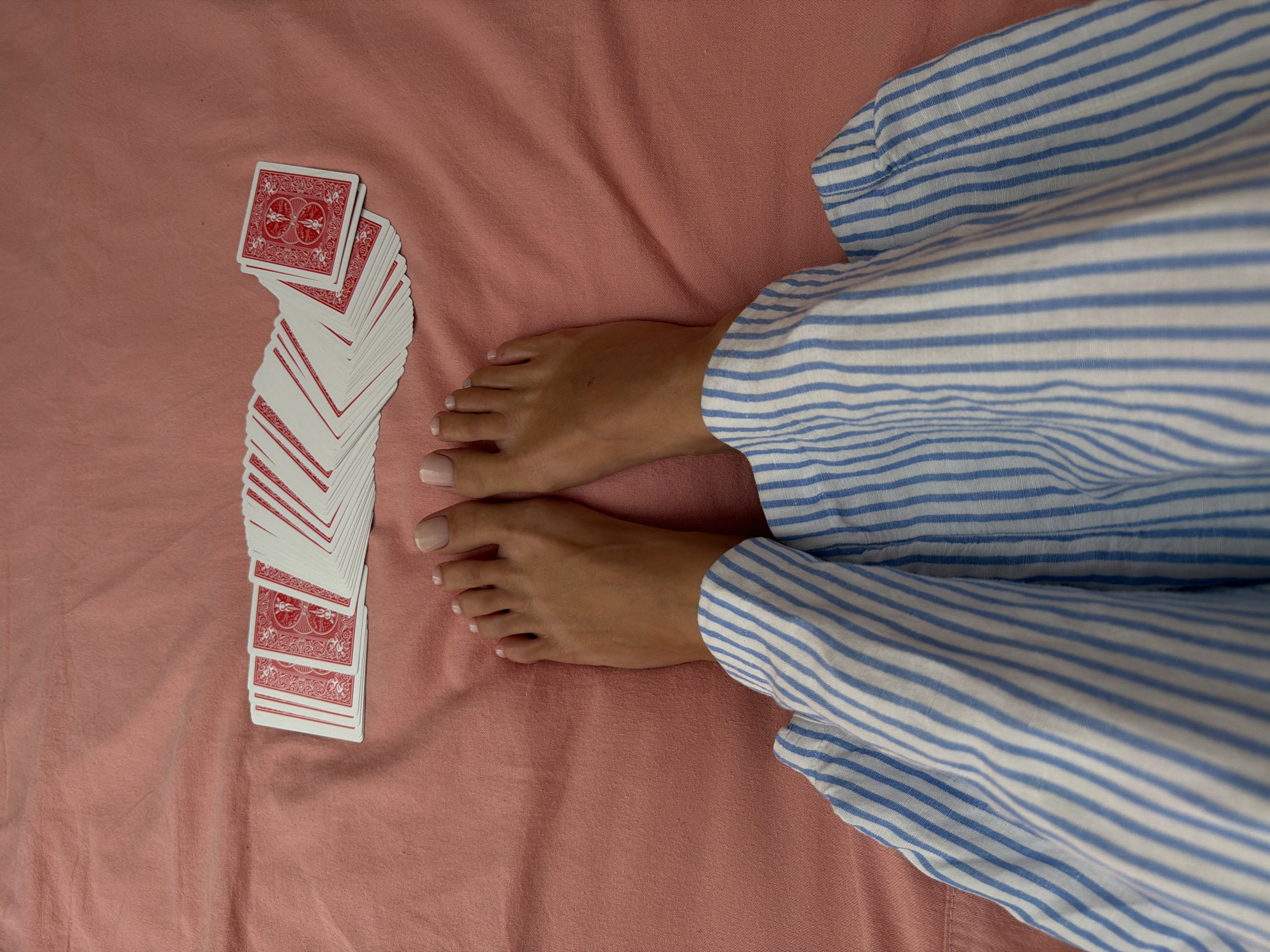The image shows a person's feet and legs resting on a pink, textured surface, likely a bed or blanket. The person is wearing blue and white striped pajama pants. To the left of the feet, a fanned-out deck of playing cards is laid out in a line, with the red backs facing up and the white sides visible. The cards are arranged in a slightly curved pattern. The scene is shot from a top-down perspective. No specific time of day, weather, or emotions are discernible. There is no visible text in the image. The location context is Mengwi, Indonesia, but this information is not visually apparent from the image itself.