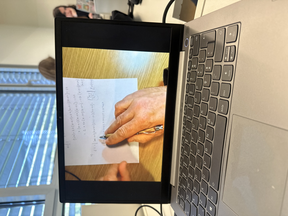 A silver-colored laptop is positioned in the foreground, resting on a white desk surface. Its screen displays a close-up image of a person's left hand, adorned with a ring on the fourth finger, holding a metallic pen and pointing at handwritten mathematical equations on a white sheet of paper. The paper lies on a light brown wooden surface. Visible equations include terms such as `P(u,v)`, `(u-0.1)`, `(0.4-0.1)(0.4-0.2)(0.4-0.3)`, `f'(t)/4T`, and the numerical result `0.7667`. The laptop features a dark grey QWERTY keyboard with a Portuguese layout. A black cable is connected to the right side of the laptop. In the blurred background, a large window fitted with horizontal venetian blinds allows natural light into the room. Two indistinct individuals are visible, seated at desks, suggesting an educational environment such as a classroom or study hall. This scene is situated within a university or educational institution in Guimarães, Portugal.