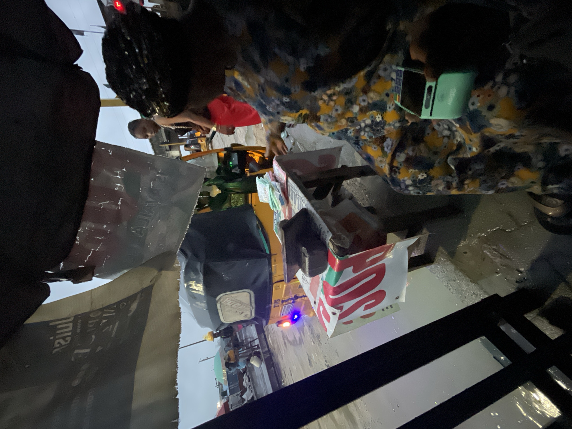 An over-the-shoulder view captures a woman in a floral-patterned dress, holding a mint-green mobile payment device, as she appears to be conducting a transaction outdoors in Lekki, Nigeria. The scene is likely a street market or roadside stall, with a yellow tricycle, a common mode of transport and vending in Nigeria, parked nearby. The tricycle's tail lights are illuminated, suggesting it's either active or recently used. A makeshift table is set up in front of the tricycle, covered with a banner displaying red text and a white background, on which a stack of cash and a heavy dark object, possibly a weight or a tool, are placed. The ground is wet, indicating recent rain or puddles, common during the rainy season in Lekki. A young woman with braided hair and a red skirt stands behind the table, partially obscured by the main subject. In the background, the overcast sky and other market stalls or street activities are visible, hinting at a bustling urban environment. The overall atmosphere is one of everyday commerce in a West African city.