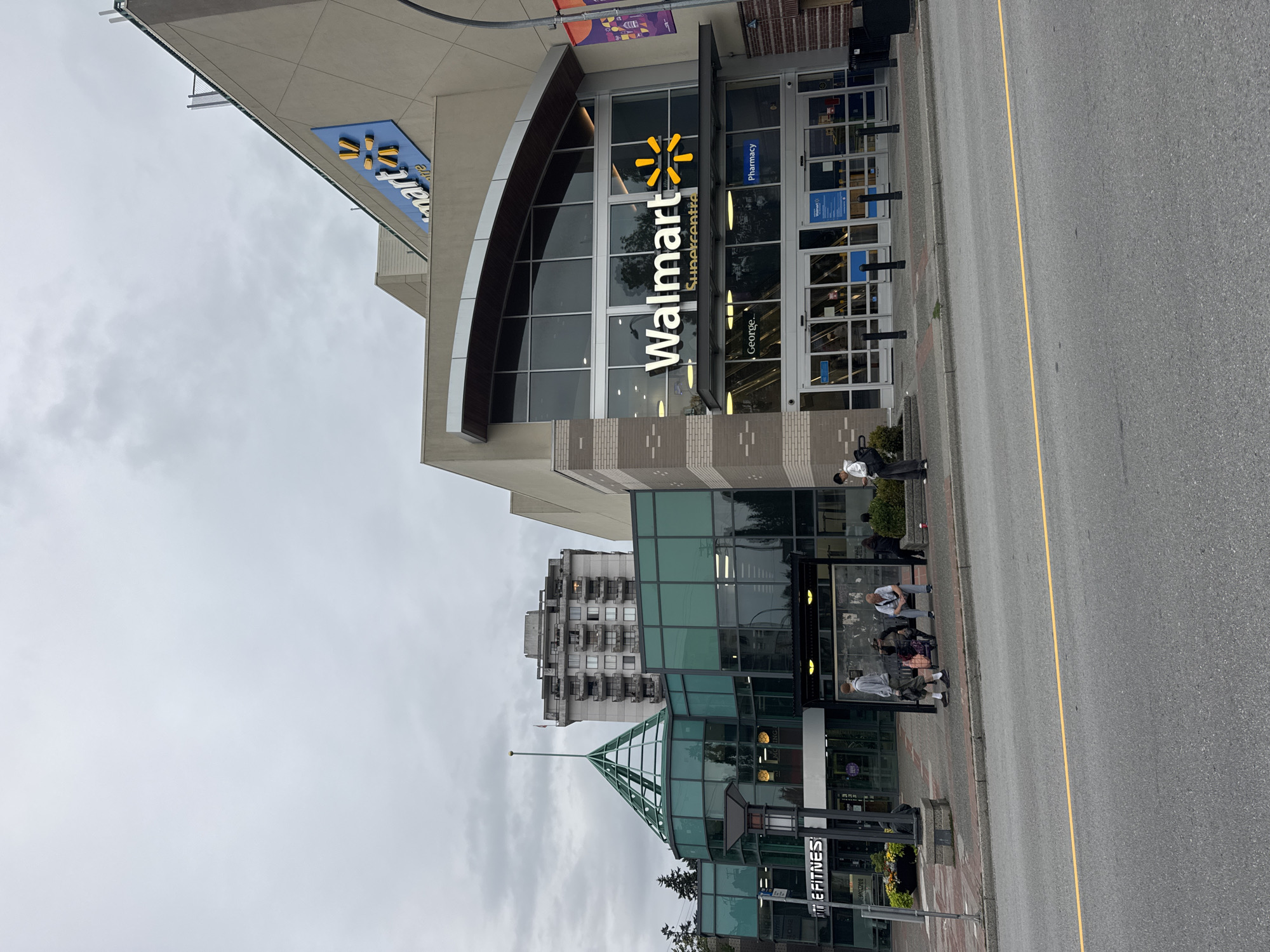 The image shows the exterior of a Walmart Supercentre and an adjacent building with a distinctive glass-domed entrance in New Westminster, Canada. The Walmart store features large glass doors, a prominent "Walmart" sign with the signature spark logo, and banners. To the left of Walmart, the adjacent building has a modern architectural design with extensive glass paneling and a green-tinted glass dome. A sign for "THE FITNESS" is visible on the left side of this building. Several people are depicted in the scene. Some are sitting at a bus stop located on the sidewalk in front of the buildings, while others are walking or standing nearby. The street in front of the buildings has asphalt pavement with a yellow line marking the lane. The sky is overcast, suggesting a cloudy day. In the background, a multi-story residential building with many balconies is visible, as are trees and parts of other structures.
