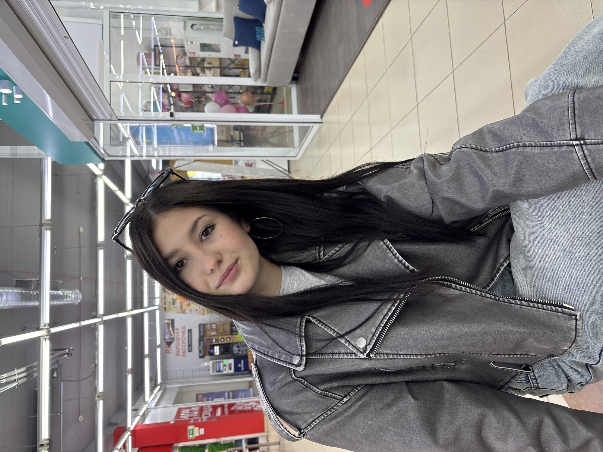 The image shows a young woman with long dark hair, wearing a gray jacket and jeans. She is positioned inside a store in Chaikovsky, Russia. The store appears to be a retail establishment with shelving visible in the background, including items like home goods and electronics. Overhead lighting is prevalent throughout the store. The woman is looking directly at the camera with a slight smile.