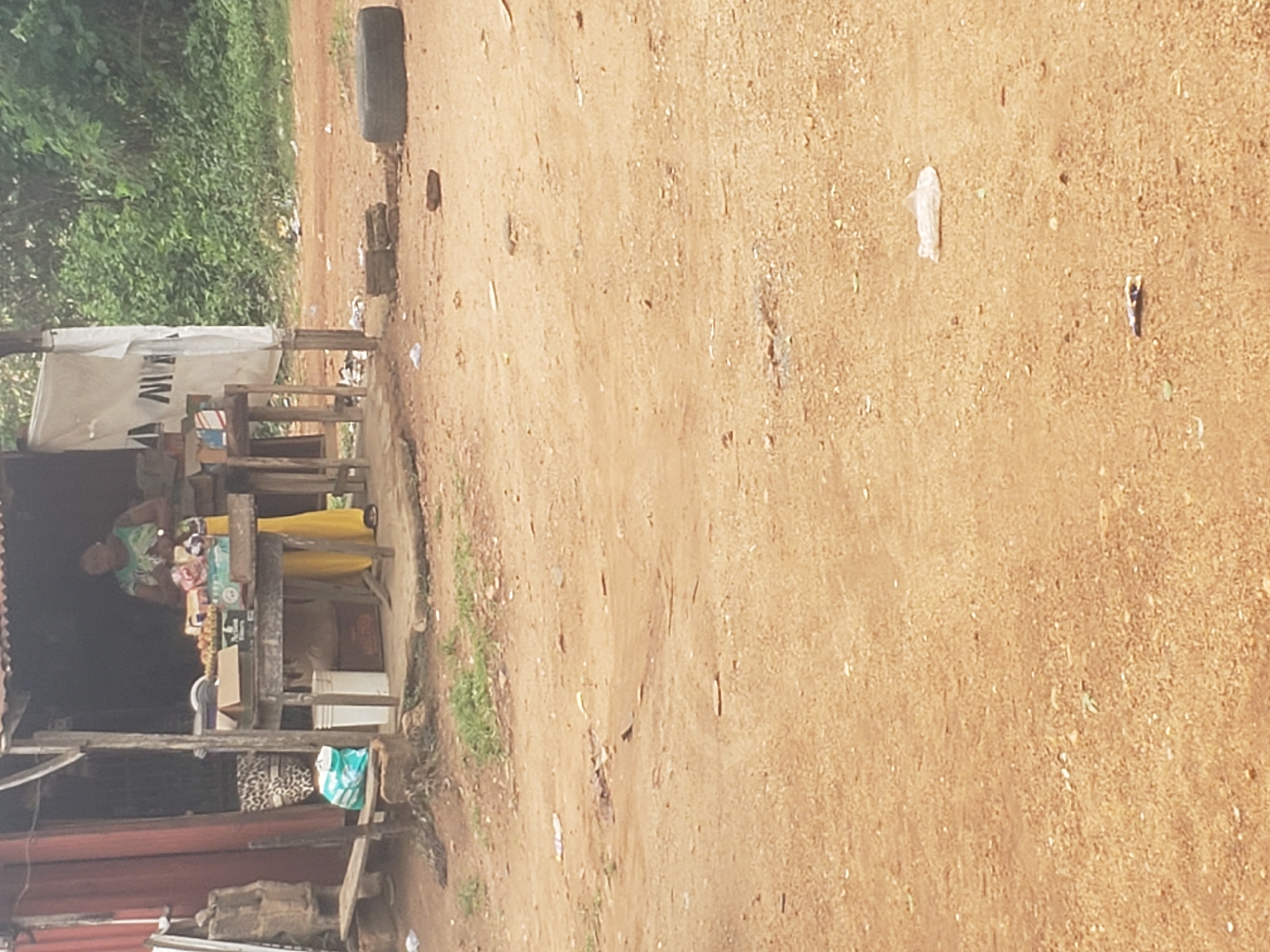 This media file captures a daytime scene of a roadside vendor stall in a rural or semi-urban setting, consistent with the Ibadan, Nigeria context. The ground is a wide, unpaved, reddish-brown dirt path or road, indicating dry, dusty conditions. Lush green foliage forms a dense backdrop on the left side of the image.
The central subject is a makeshift vendor stall constructed from wooden planks and poles, topped with a corrugated metal roof and partially covered by a white tarp. Behind the counter, a woman, likely the vendor, is visible. She wears a light-colored, possibly green or patterned top, and a bright yellow skirt or wrapper, and appears to be attending to the goods on display.
The stall's counter holds various items for sale, including packaged snacks, beverages, and other sundry goods. Several boxes and containers are arranged on and below the counter. A blue and white patterned bag and another patterned bag are visible near the base of the stall structure.
On the dusty ground, scattered litter includes a distinct white plastic bag and other small pieces of debris. Further down the path, a black car tire rests on the ground.
The visible text "AW" can be seen on the white tarp covering part of the stall. Other text on product packaging is too small or blurry to read. The overall scene depicts a typical local commerce activity.
