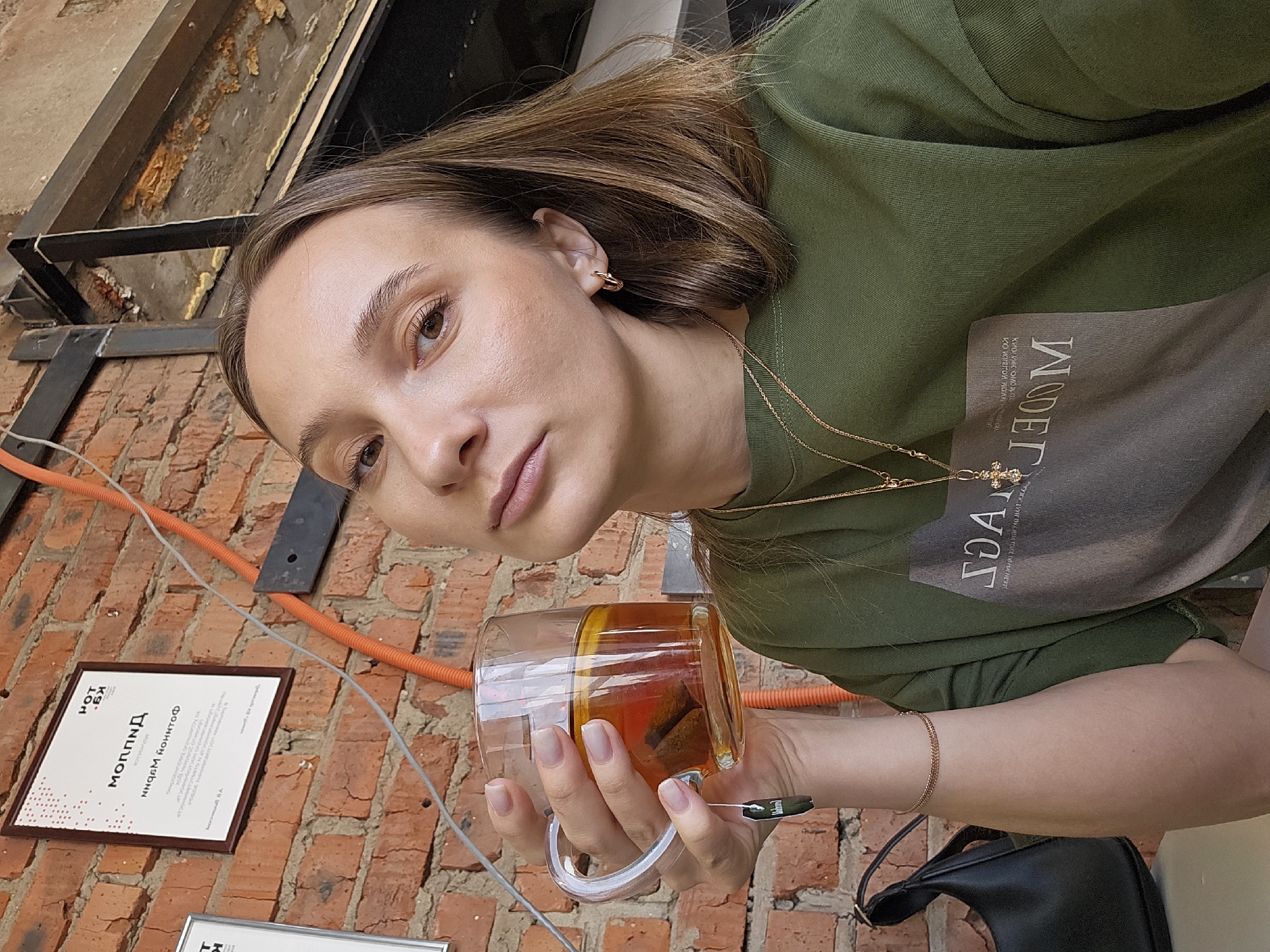 A woman with brown hair and fair skin is holding a clear glass mug filled with a dark liquid, possibly tea, in what appears to be an outdoor setting in Chaikovsky, Russia. She is wearing a green top with the word "MODEL" printed on it. The mug contains a slice of lemon and a tea bag. A thin gold necklace with a cross pendant is visible around her neck, along with a gold bracelet on her wrist. A black purse is partially visible at the bottom right.
The background features a red brick wall with an orange electrical conduit and a framed document with Russian text. The document has "МОЛПИД" in larger letters at the top. A metal beam structure is visible above the woman's head.
