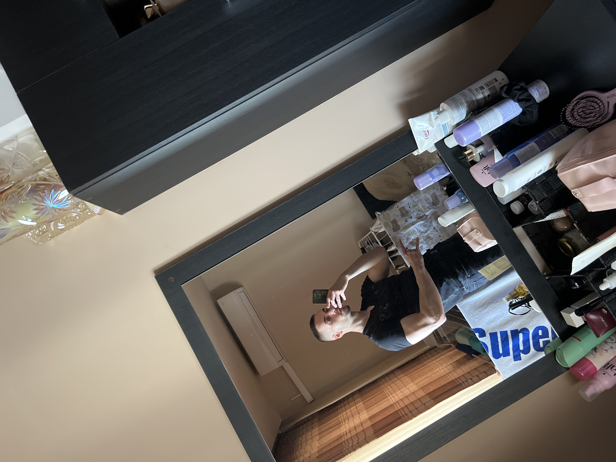 A mirror reflects a person taking a selfie, along with elements of a room. The person is facing the mirror, holding a phone in one hand, and making a gesture with the other. The reflection also shows an air conditioner, curtains, and part of a towel with the word "Super". The immediate surroundings of the mirror include toiletries, makeup, and other items on a shelf. The overall setting is indoors, likely a bathroom or dressing area. Burgas, Bulgaria is not discernible.