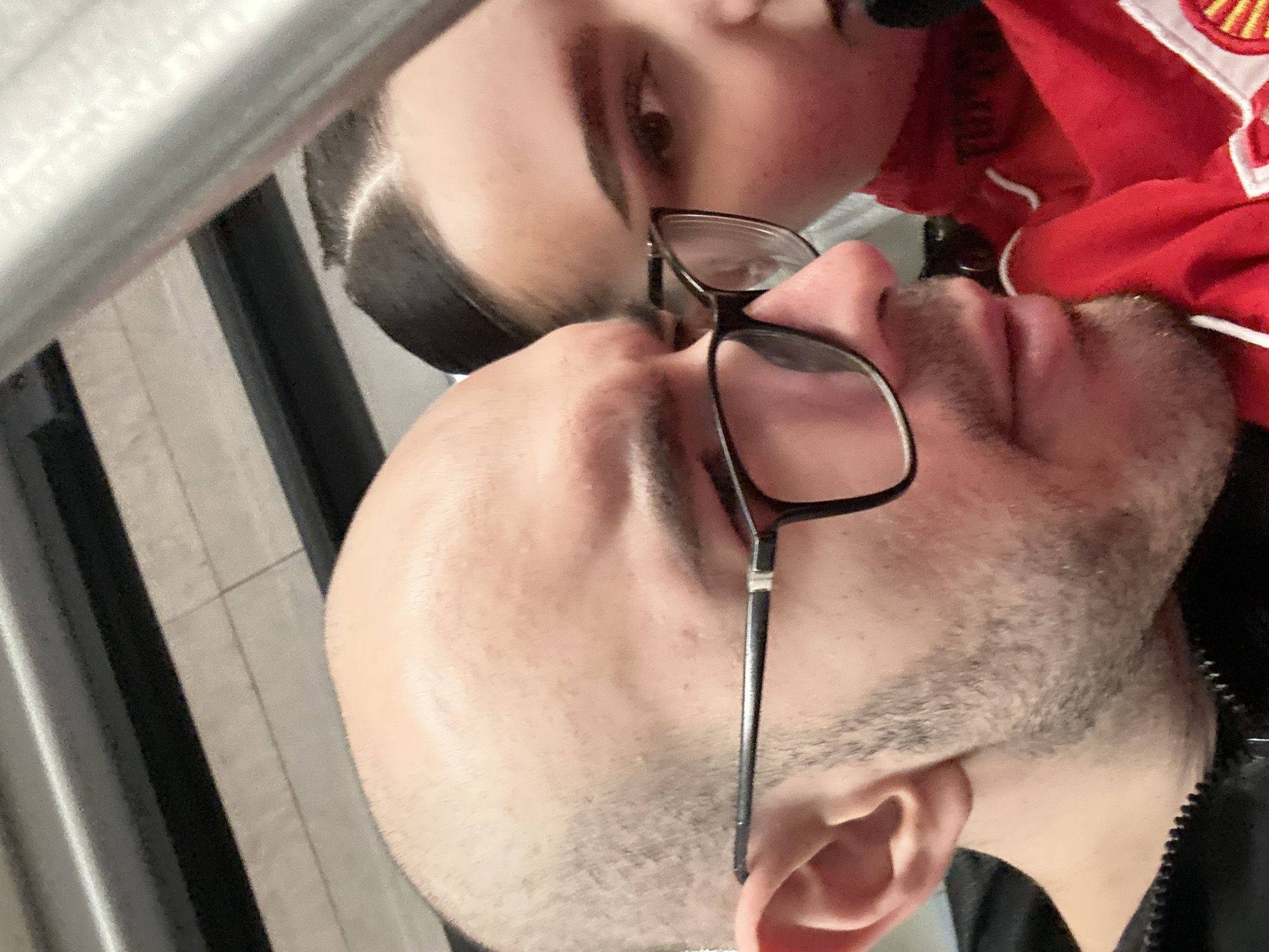 A selfie-style image presents two individuals in close proximity. The person in the foreground wears glasses and has visible facial hair. The other person is partially visible, wearing a red garment with white accents and an emblem. The setting appears to be indoors, with vertical striped elements in the background. The location is not discernible from the visual information provided.