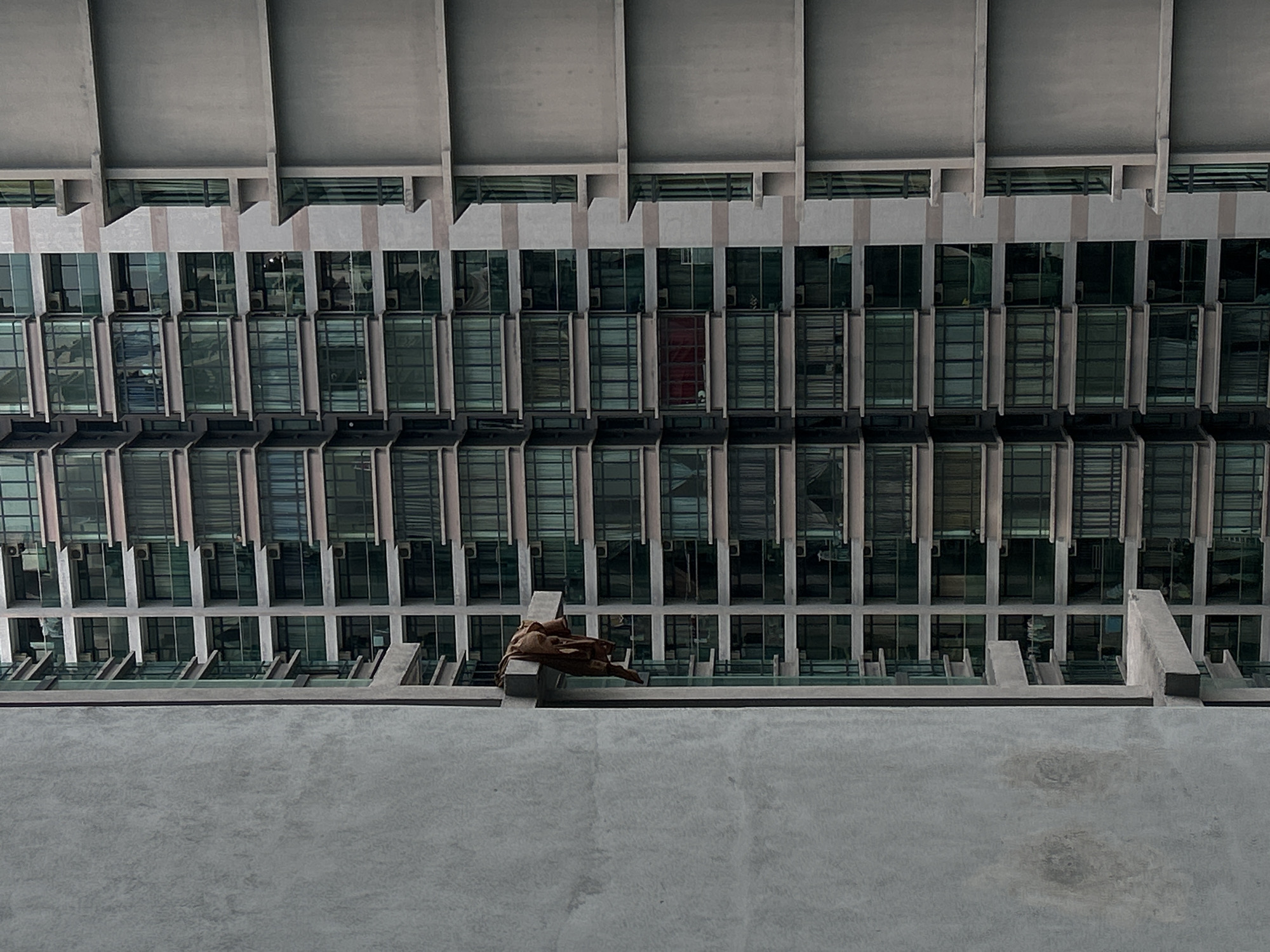 A low-angle shot of a building facade in Kuala Lumpur, Malaysia. The facade consists of repeated rows of windows, each with a dark-tinted glass surface. Vertical concrete columns separate each window unit. The top of the building features overhanging concrete slabs with recessed lighting or ventilation elements. A sculpture in brown tones is visible at the lower portion of the frame, on a ledge. The sculpture is possibly a representation of a figure. A large concrete surface occupies the lower third of the frame, appearing to be part of a wall or parapet from which the photograph was taken. The overall palette is muted and grayscale, adding to a stark, architectural feel.