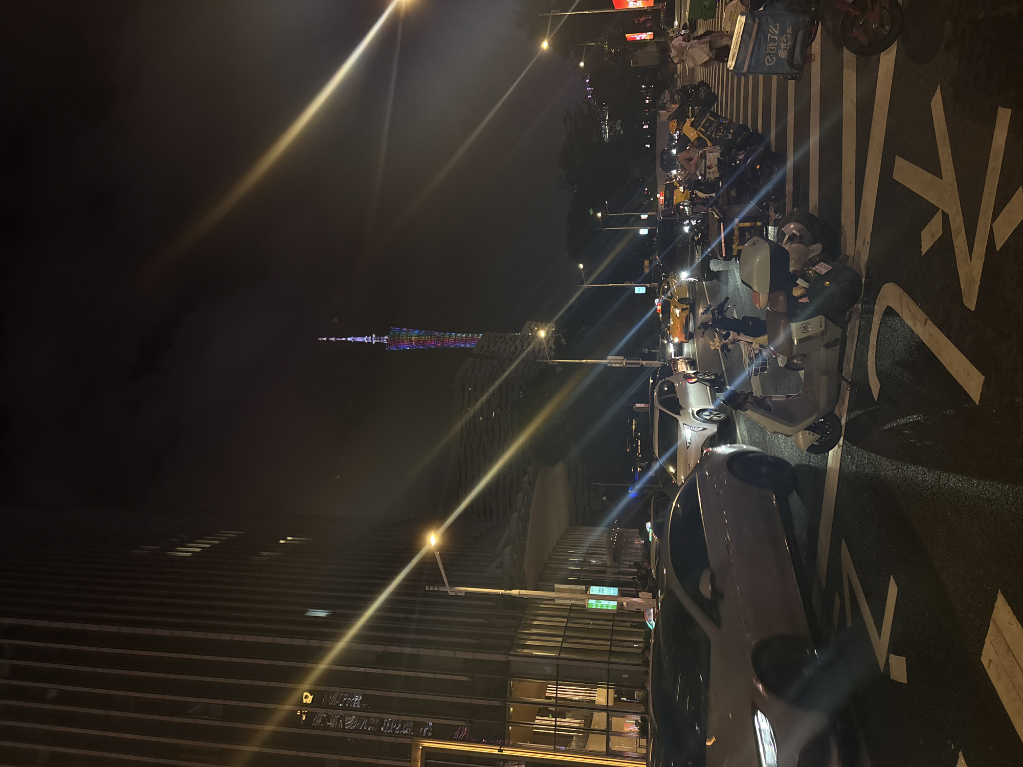 A nighttime, street-level, medium-shot photograph captures a busy intersection in Tianhe District, China. In the foreground, a white electric scooter is parked on a crosswalk, its handlebars pointed to the left. To its right, a delivery person wearing a white helmet and carrying a large blue delivery box on their back is visible. Further back, several more scooters are parked. Cars, including a white sedan, a silver SUV, and a yellow taxi, are present in the traffic lanes. The background shows a modern skyscraper with vertical lines of windows and a brightly lit, colorful tower in the distance, likely the Canton Tower, illuminated against the dark, slightly hazy sky. Streetlights cast a yellowish glow on the scene, and diagonal light streaks, characteristic of lens flare, are prominent across the image.
