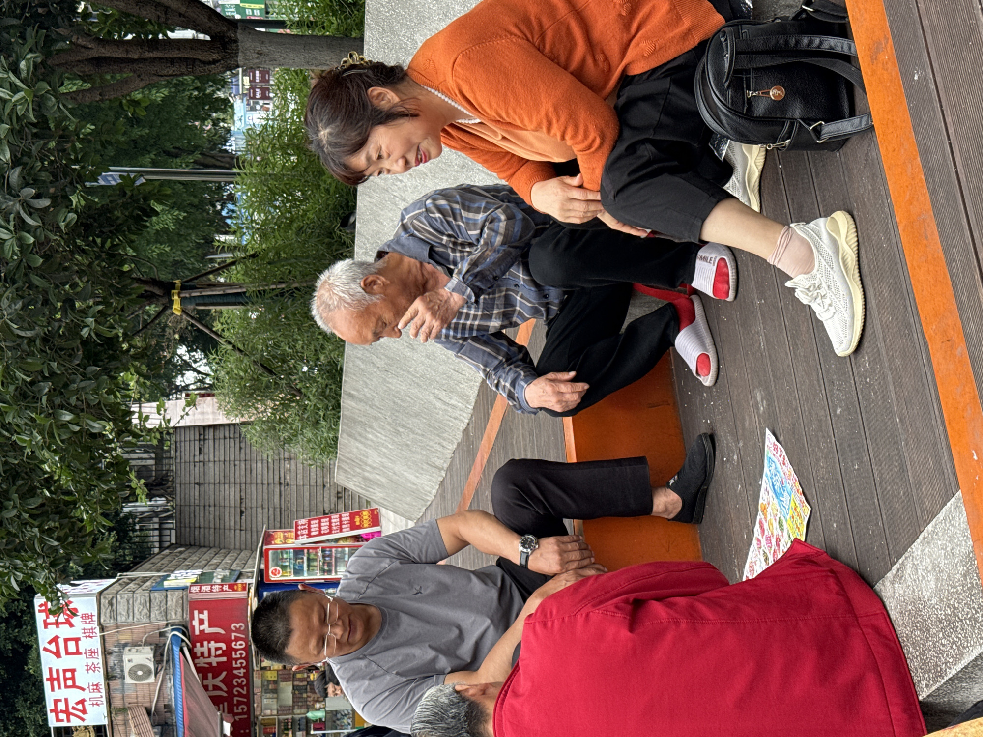 Here is a description of the image:
**Location:** Nan'an District, China. This is inferred from the visible Chinese signage.
**Scene Description:** The image shows a street scene where several people are interacting.
**People:**
* Two older people, a man and a woman, are seated on a raised wooden platform. The woman is wearing an orange sweater and black pants. The man is wearing a plaid shirt and black pants. The woman appears to be attending to the man, who is holding his neck or head.
* Two additional men are on the ground. One is wearing a grey shirt and black pants, and appears to be looking at or tending to the other, who is lying on his back and partially covered by a red blanket.
**Objects:**
* A black bag is next to the woman.
* A flyer or printed material is on the wooden platform.
* Various storefronts and signage are visible in the background, suggesting a commercial area.
* An air conditioning unit is attached to one of the buildings.
**Actions and Interactions:**
* The woman appears to be attending to the man seated next to her.
* The man in the grey shirt appears to be tending to the man lying on the ground.
**Environment:**
* The scene is outdoors, likely on a public street.
* The presence of trees and buildings indicates an urban or semi-urban setting.
* The wooden platform suggests a designated seating area or public space.