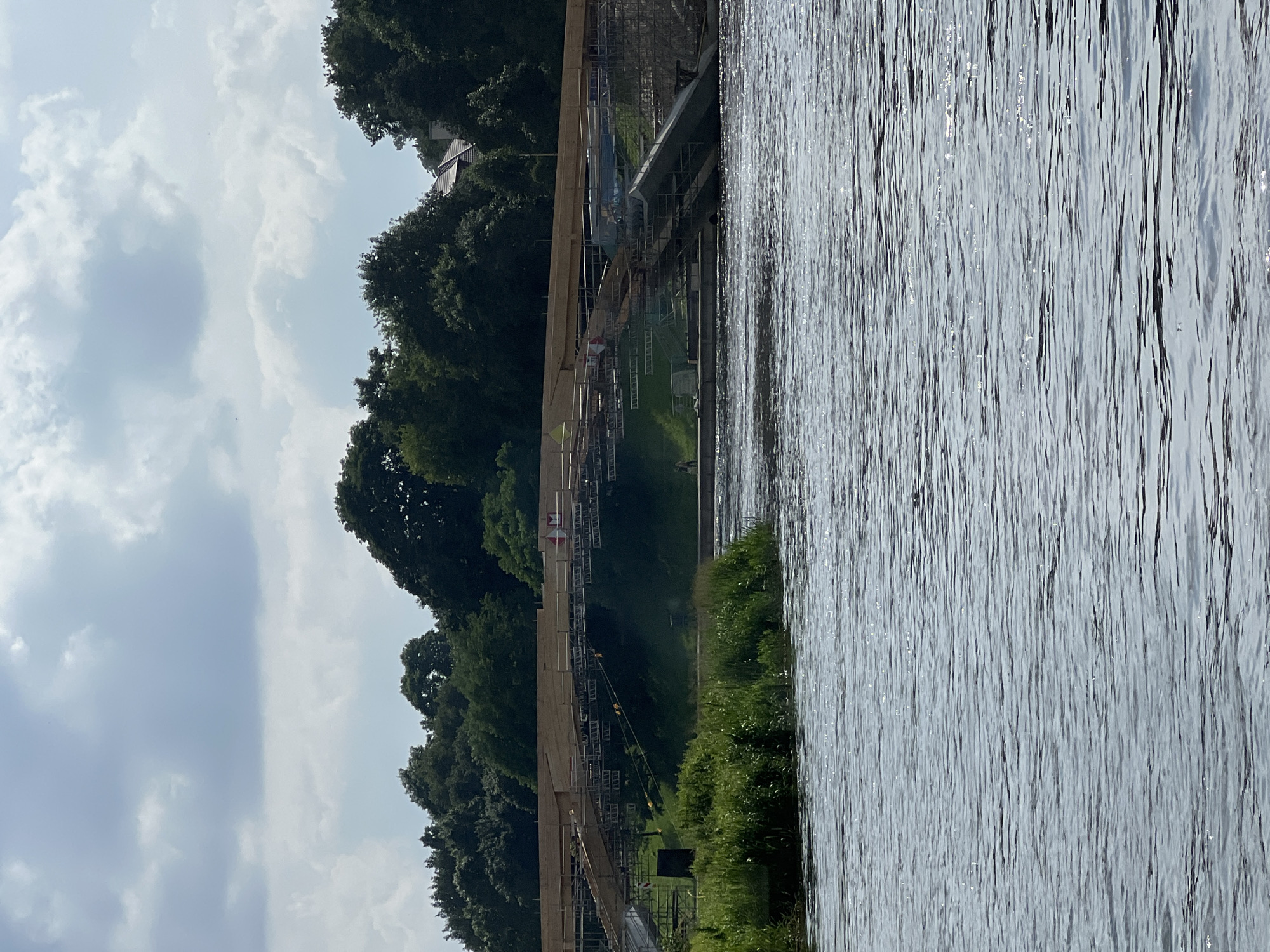 The image shows a river with a bridge under construction. The bridge is made of wood and metal scaffolding. There are trees and greenery on the river banks. The sky is cloudy. There is one person visible on the bridge, and another person on the river bank. The location is likely Vilnius, Lithuania, as this is the site of a bridge renovation project on the Neris River near the Green Bridge.