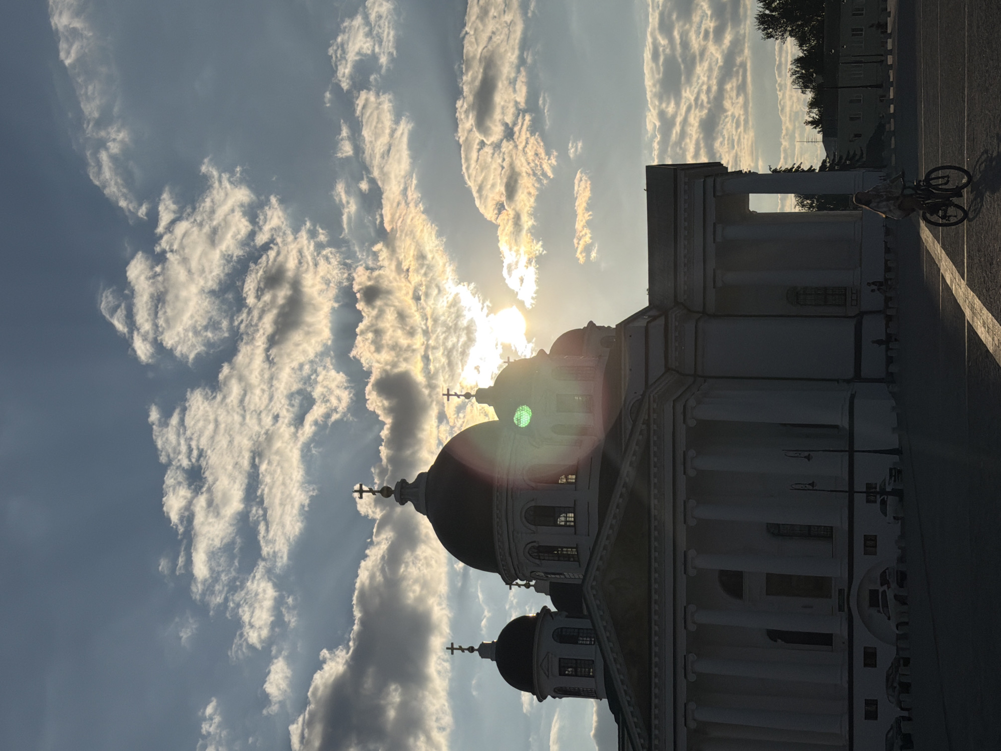 The image captures the Arzamas Cathedral in Arzamas, Russia, during sunset. The sun is low in the sky, casting long shadows and creating a dramatic backlight effect that silvers the clouds. The cathedral, with its prominent domes and columns, is a majestic presence against the vibrant sky. In the foreground, a young woman is seen riding a bicycle. The scene is peaceful and serene, with a sense of tranquility as the day comes to a close.