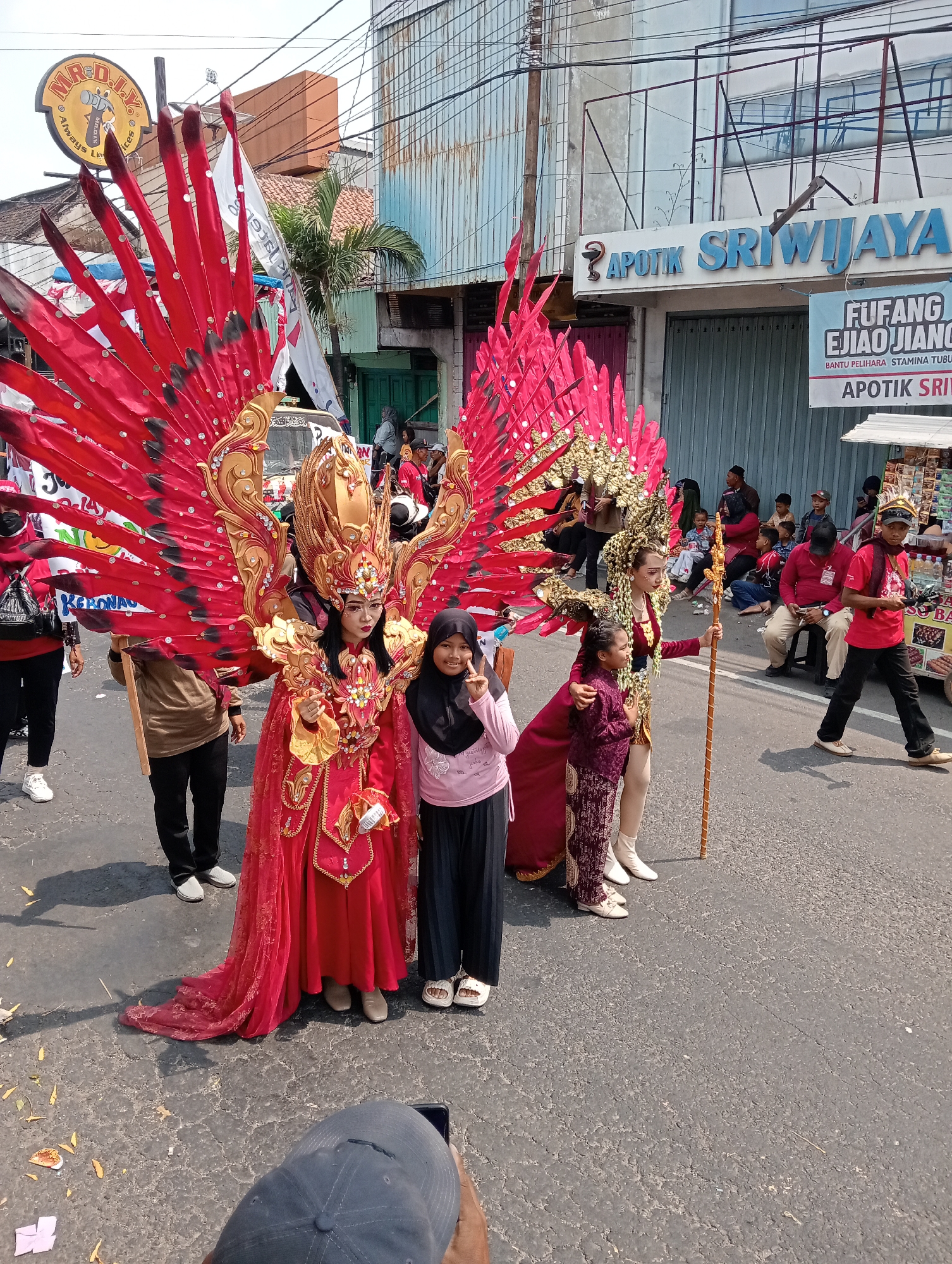The image captures a street scene in Demak, Indonesia, likely during a parade or cultural event. In the foreground, a person dressed in an elaborate red and gold costume with large, feathered wings stands prominently. To their left, another individual in a similar costume is partially visible. To the right of the main costumed figure, a young girl in a pink shirt and black pants poses for the camera, making a peace sign. Further to the right, another young person is seen in a red shirt and black pants. A group of spectators, including children and adults, are seated and standing along the roadside, observing the event. In the background, buildings line the street, with one featuring a sign for "APOTIK SRIWIJAYA." The street is paved and appears to be in a moderately urban setting. The overall atmosphere suggests a festive occasion.