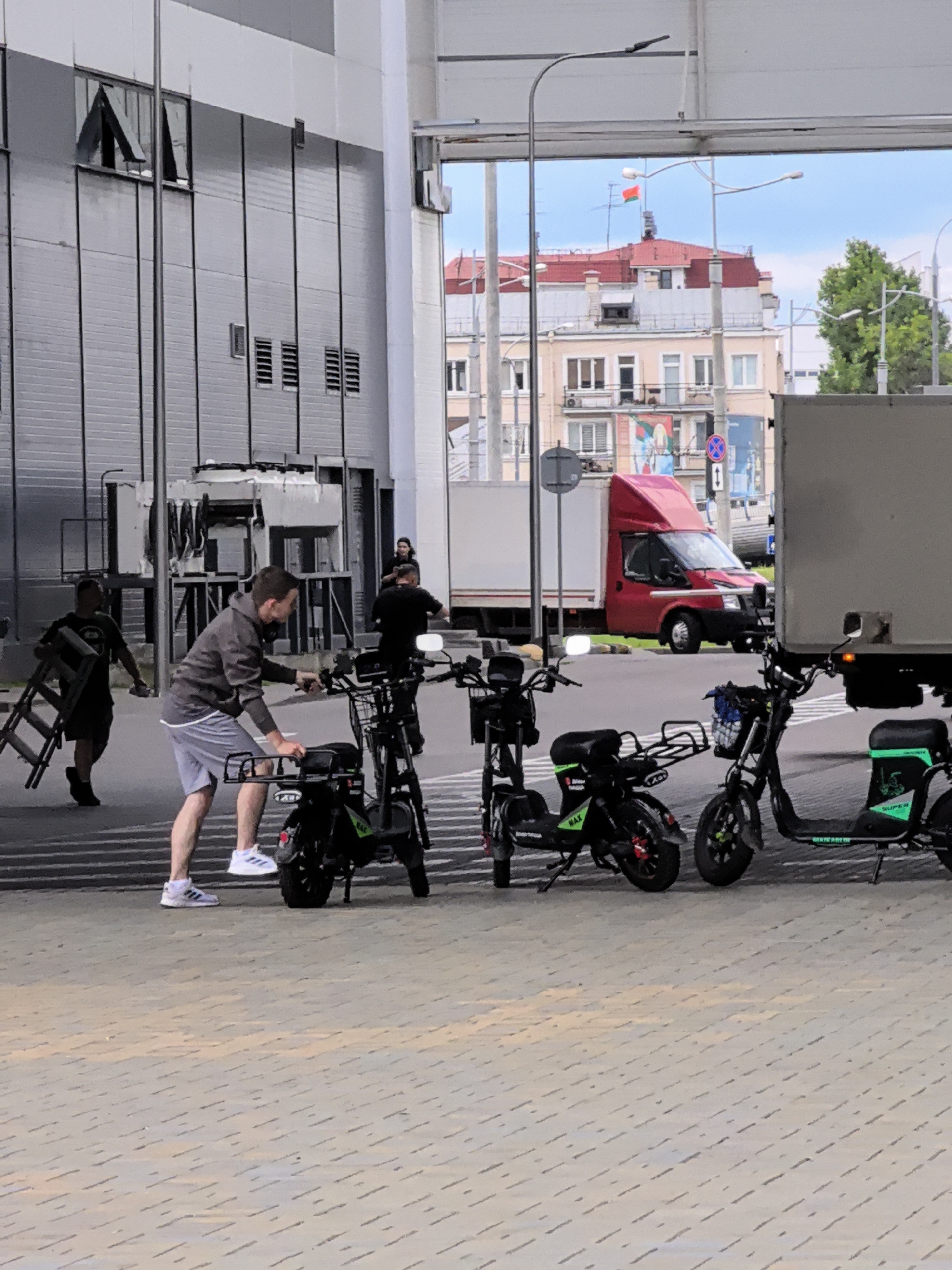 In a city environment, under overcast skies, a man in a gray hoodie and gray shorts stands next to a black electric scooter, adjusting its seat. He wears white athletic shoes. To his left, another man in a dark shirt and shorts carries a silver folding ladder. In the background, several other black electric scooters are parked in a row. A red van and a large gray truck are visible behind the scooters. A modern building with dark gray siding and windows forms the backdrop to the left. In the distance, a multi-story building with a red roof and a small flag is visible. The scene appears to be in an urban outdoor area, possibly a loading dock or a public street.