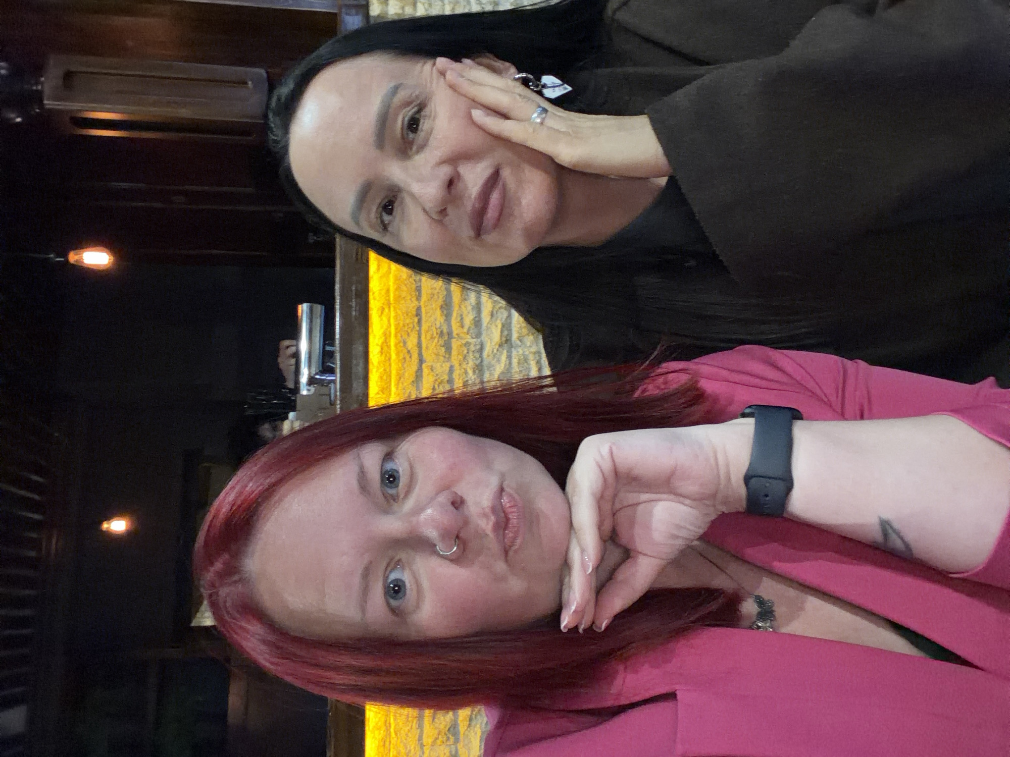 Two women are posing for a selfie. The woman in the foreground has red hair, blue eyes, a nose ring, and is wearing a pink jacket and a black smartwatch. Her right hand is resting on her cheek. The woman in the background has long black hair and is wearing a brown jacket. Her left hand is touching her cheek. The background appears to be a dimly lit indoor setting, possibly a bar or restaurant, with a yellow decorative wall and some blurred lighting fixtures. The exact location in the city of Chaikovsky, Russia, is not discernible from the image.