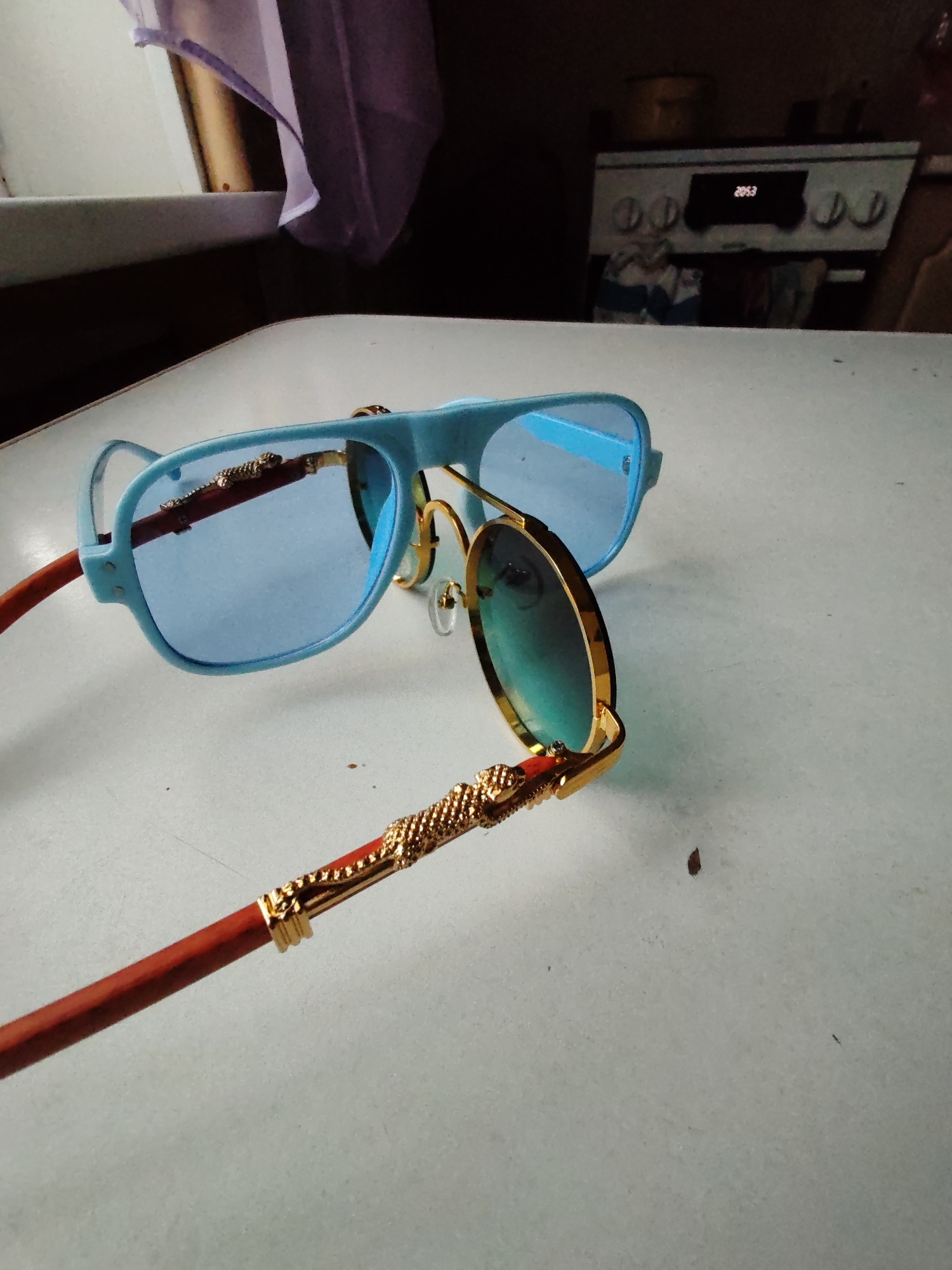 The image shows a pair of blue-rimmed sunglasses with blue lenses and gold accents on a white surface. One lens is in focus, showing a reflection of a gold, ornate temple arm. The other pair of sunglasses is slightly out of focus, with a blue frame and blue lenses. The background is blurred but suggests an indoor setting with a kitchen appliance visible. There is no indication of the city of Чухлэм, Russia.