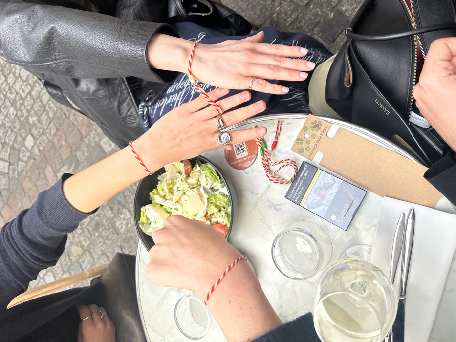 A top-down view depicts a forensic scene at an outdoor cafe in Paris, France. Three individuals are partially visible, primarily represented by their arms and hands around a white marble-patterned round table with a metallic edge.
On the left, one person, wearing a dark long-sleeved top, has their right forearm and hand on the table. This hand wears a red and white twisted string bracelet and holds a spoon over a black bowl containing a salad of lettuce, cherry tomatoes, and sliced hard-boiled eggs.
In the center-right, another individual's left forearm and hands are visible. This person wears a black leather jacket over dark blue patterned pants. Their left wrist also bears a red and white twisted string bracelet. Rings adorn two fingers of this hand: a large, intricate silver ring on the ring finger and a gold-toned band on the middle finger.
The table holds several objects:
* A black bowl of salad.
* Two empty clear glasses, possibly wine or water tumblers.
* A white folded napkin with two silver pieces of cutlery.
* A packet of "SOBRANIE" cigarettes.
* A brown cardboard card with a green four-leaf clover design and a red ladybug, bearing the Cyrillic text "Честита Баба Марта" (Happy Baba Marta).
* An additional red and white twisted string bracelet, accented with green beads, lies coiled near the card.
* A red QR code sticker is affixed to the table surface.
In the bottom right, a third person's hand reaches into an open black leather "LOEWE" branded handbag, with a gold-toned ring visible on one finger.
The environment indicates an outdoor setting with a dark cobblestone pavement visible beneath the table. A wooden chair back is partially visible on the far left.