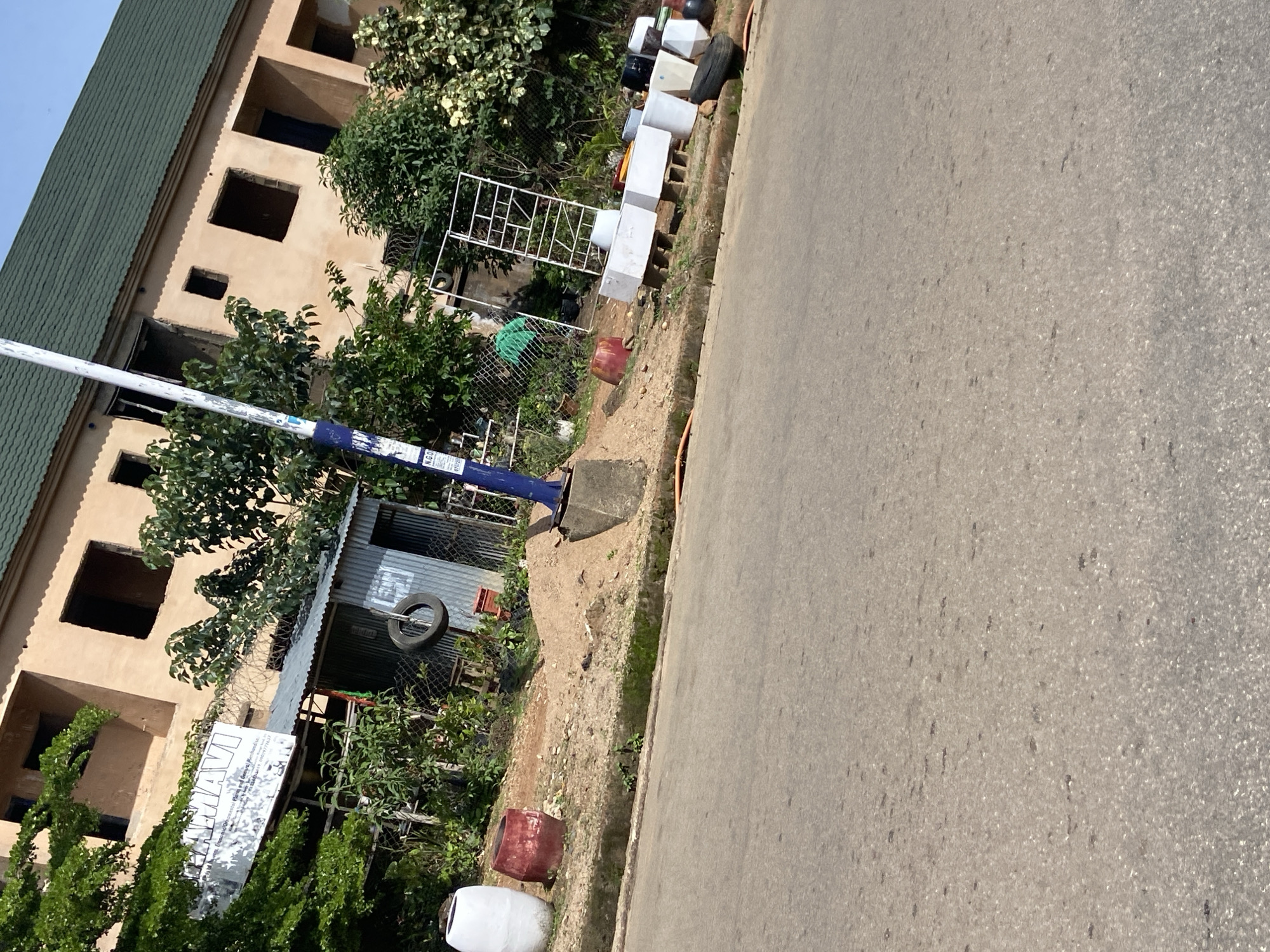 The image shows a street scene in Jos, Nigeria, with a tilted perspective. In the foreground, a paved road is visible, with a gravelly shoulder bordering it. Along the shoulder, there are several objects, including large white and red cylindrical containers, a metal rack, and what appear to be concrete planters of various sizes and colors. A discarded tire is also present.
In the mid-ground, a small corrugated metal structure stands, resembling a makeshift stall or shed, with a tire hanging from its side. Behind this structure, a chain-link fence separates the area from a building. The building itself has a green, sloped roof and a beige, unfinished facade with dark, rectangular openings that appear to be windows or doorways without frames. Lush green trees and foliage are abundant, partially obscuring the building and adding to the natural scenery.
A prominent blue and white striped pole, possibly a traffic sign or utility pole, stands vertically in the scene, leaning slightly. A white banner with text is attached to a pole in front of the building, suggesting some form of establishment or signage. The sky is visible at the top of the image, appearing bright and suggesting daytime. The overall impression is of a lively street environment with a mix of urban infrastructure and natural elements.