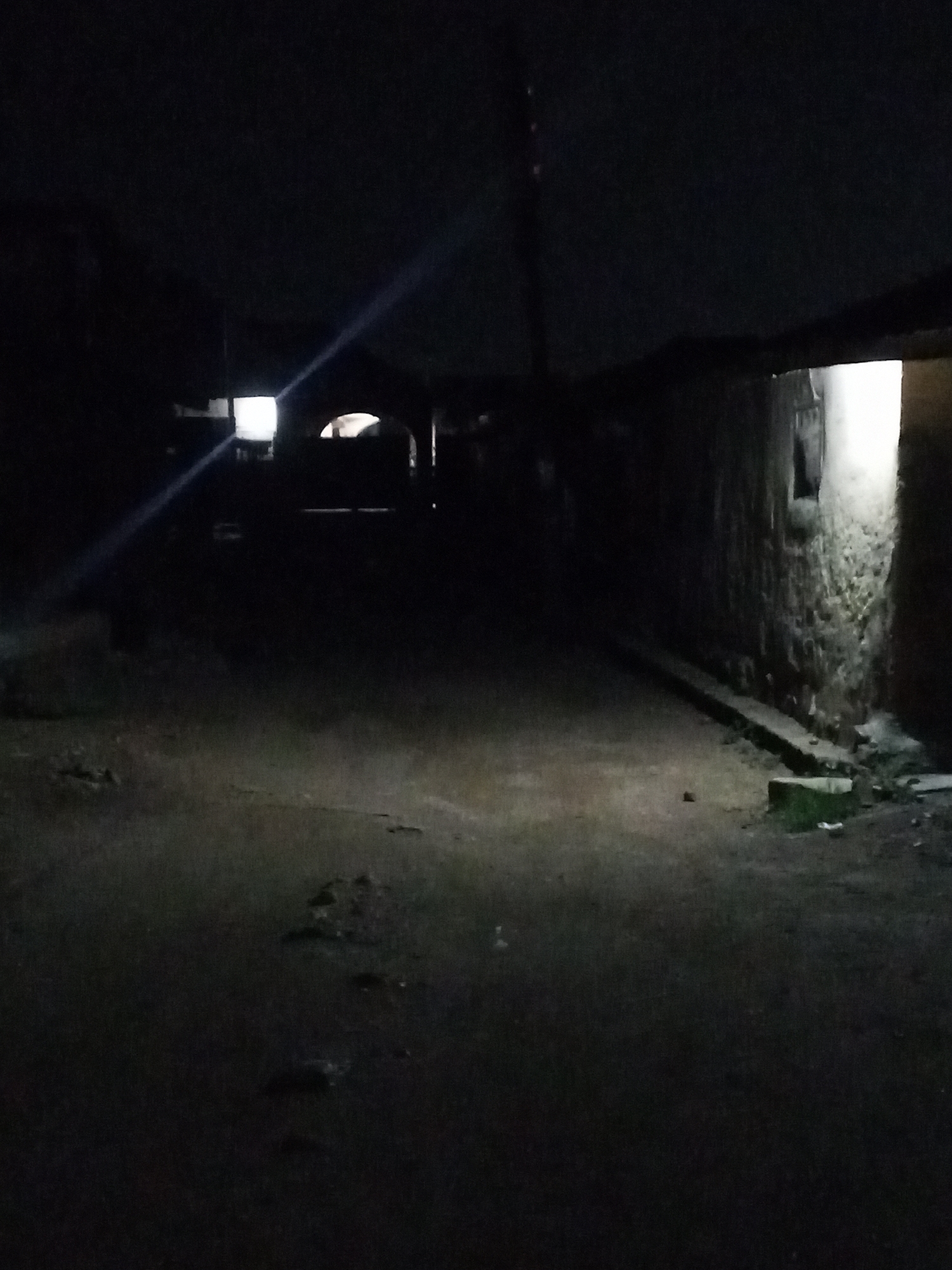 A street in Jos, Nigeria, at night. The street is dark, with minimal lighting coming from buildings. The road surface appears uneven. To the right, a structure with a window emits light. In the distance, an archway is dimly lit. A light pole stands towards the horizon. No individuals are discernible.