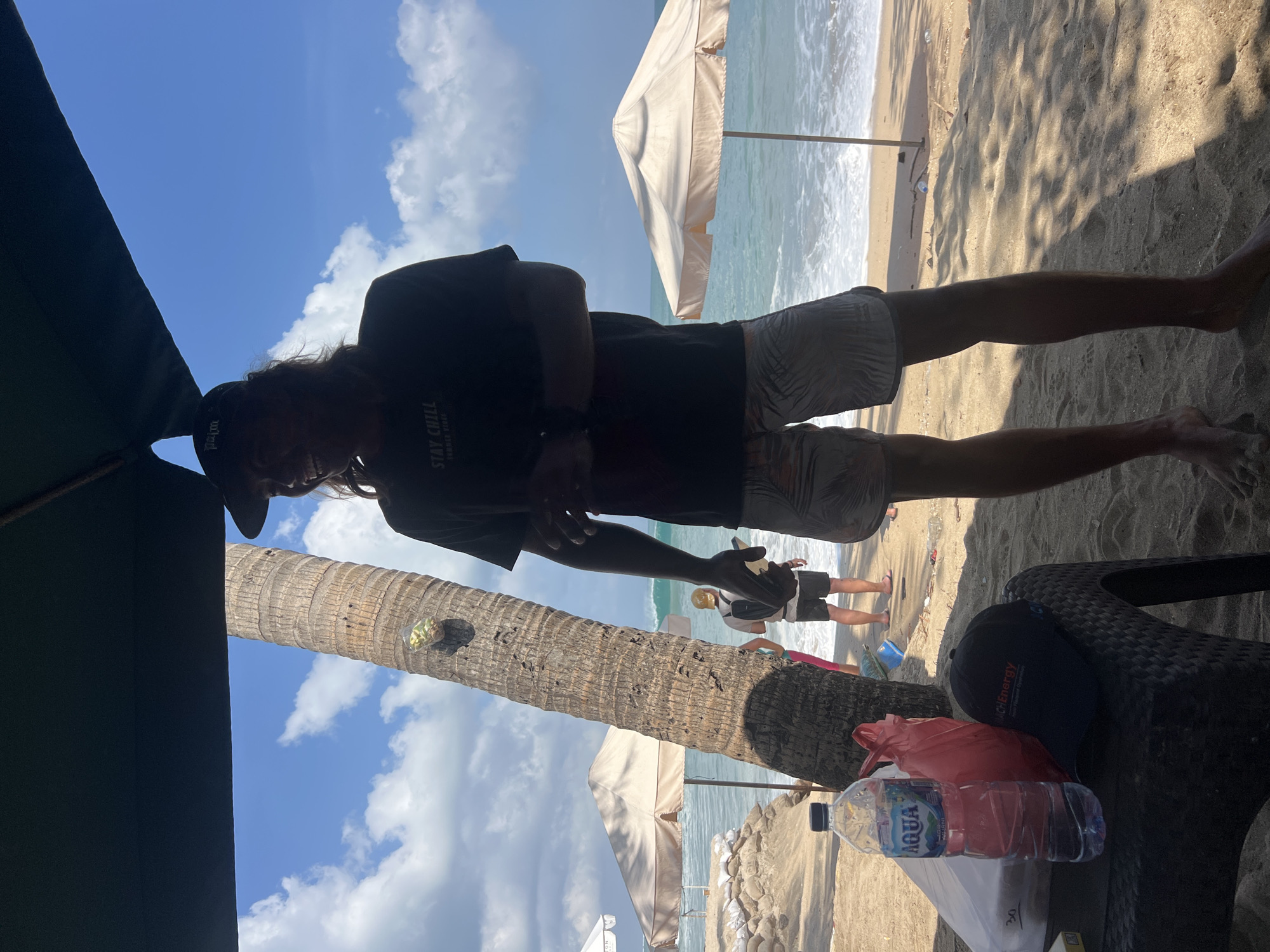 A man stands on a sandy beach in Kuta, Indonesia, under the shade of a dark umbrella. He is wearing a black t-shirt that reads "STAY CHILL BEACH VIBES" and patterned shorts. In his hand, he holds a small object, possibly a phone or camera. In the foreground, a black plastic table holds a bottle of Aqua water, a red plastic bag, and a black baseball cap with "Mach Energy" embroidered on it. Behind him, a palm tree with a cluster of items attached to its trunk stands tall. The ocean is visible in the background, with waves breaking on the shore and two beige beach umbrellas set up further down the beach. Other people can be seen walking along the shoreline. The sky is a clear blue with scattered white clouds.