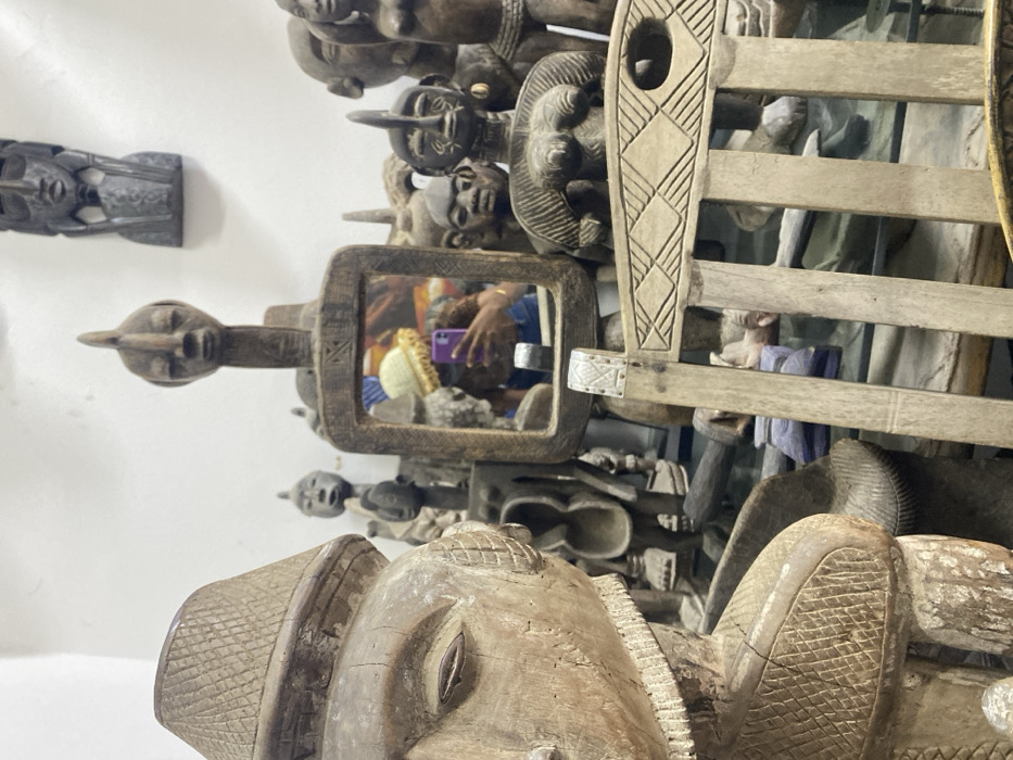 The image depicts an interior space, identified as being in Alaguntan, Nigeria, densely populated with traditional carved wooden artifacts. In the left foreground, a large, light-brown, weathered wooden sculpture of a human figure, possibly male, is prominently positioned. This figure wears a patterned, wide-brimmed hat, and features a detailed, carved left eye.
Centrally in the mid-ground, a rectangular, intricately carved wooden mirror reflects a person holding a purple-cased smartphone. The individual in the reflection possesses dark skin, wears a light-colored, possibly yellow, knitted hat with a ruffled brim, and a dark top. This reflection indicates the photographer.
Surrounding the central mirror and extending into the background are dozens of additional dark-toned wooden sculptures. These include various masks, busts, and full figures, many exhibiting distinct human or stylized features. Several darker wooden masks are mounted directly onto a plain white wall in the upper background. To the right side of the mid-ground, a section of a light-colored carved wooden chair, featuring geometric patterns on its backrest, is partially visible. The overall environment suggests a collection or retail display of traditional African art.