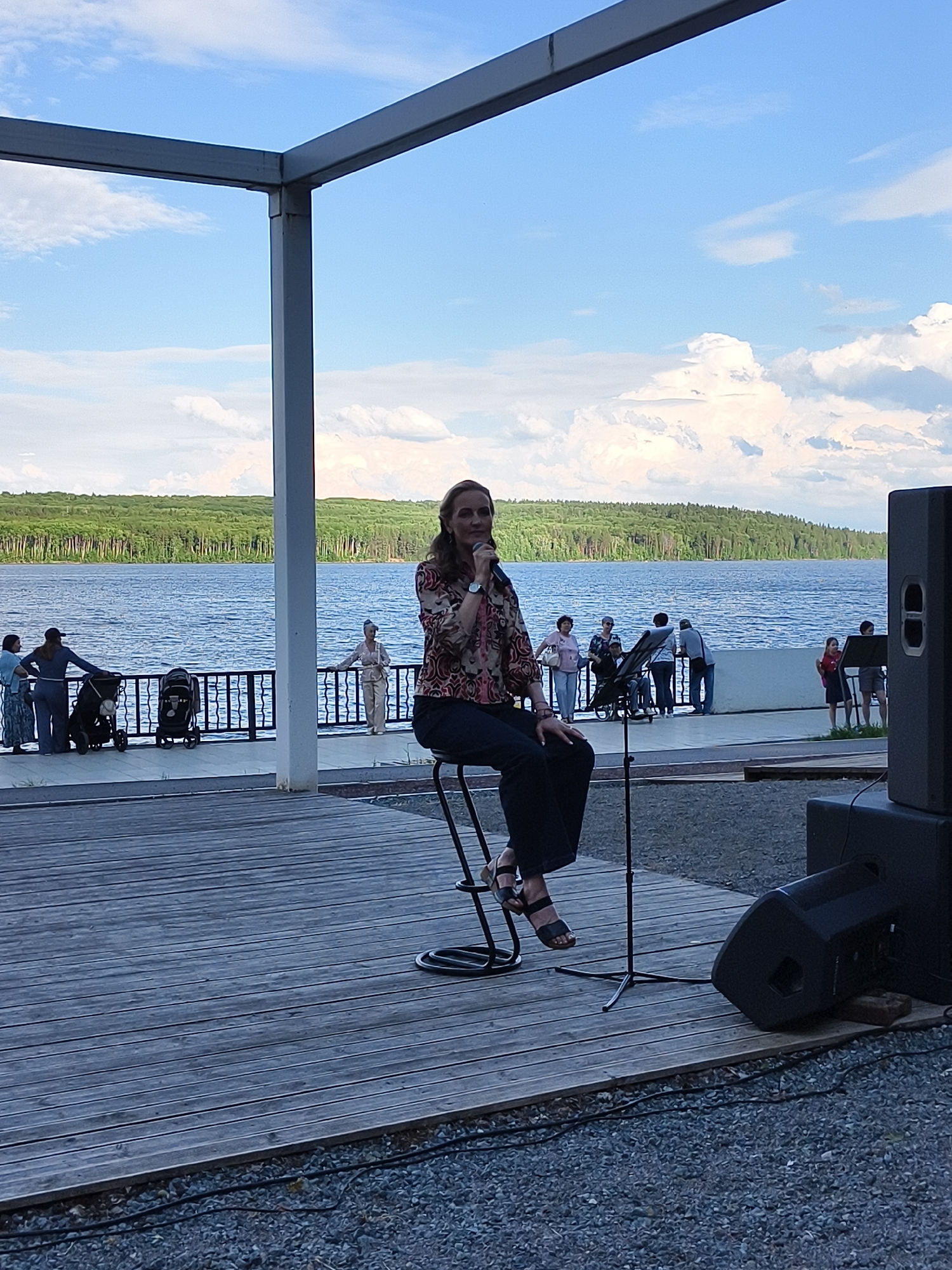 A woman is seated on a high stool, holding a microphone and speaking into it. She is wearing a patterned shirt and dark pants, and black sandals. The stool has a metal frame and is placed on a wooden deck. Behind her, a body of water is visible, with a line of trees on the far shore. Several people are standing along a railing near the water. The sky is blue with scattered clouds. To the right of the woman, there is a large black speaker. A metal pole with a circular base supports the microphone. The overall setting appears to be an outdoor venue, possibly near a lake or river.