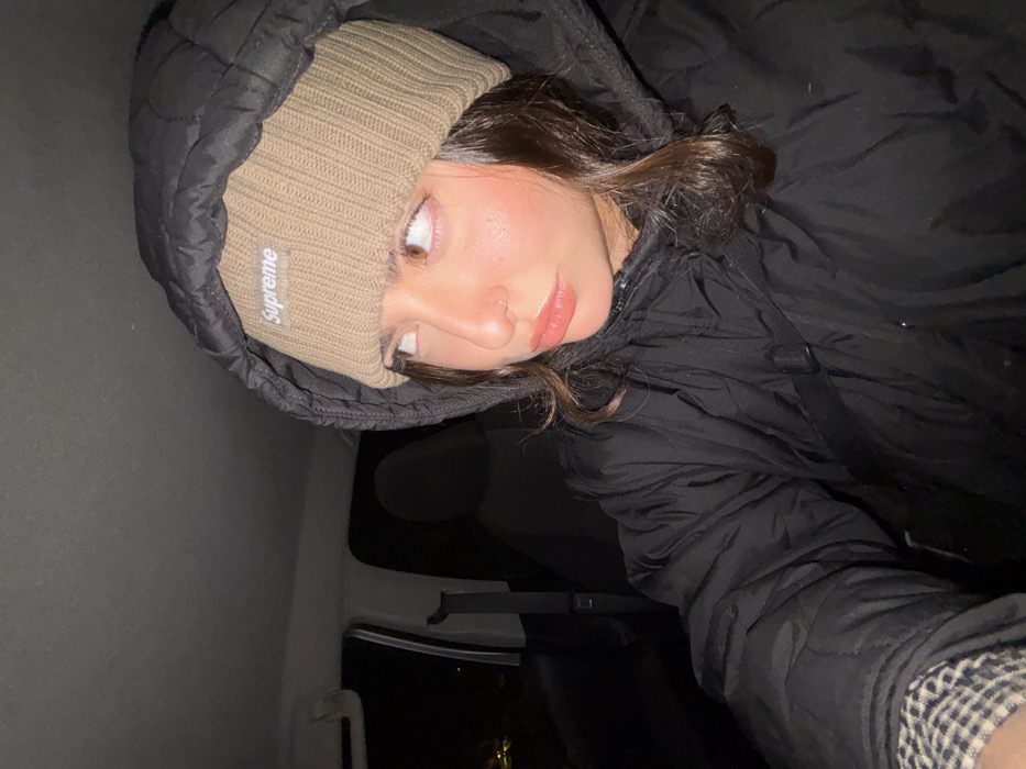 A young woman in a black hooded jacket and beige "Supreme" beanie gazes off-camera. Her lips are glossy, and she has a nose ring. The scene is dimly lit, possibly inside a vehicle, with a dark interior visible in the background. The image is a close-up, selfie-style shot angled downward.