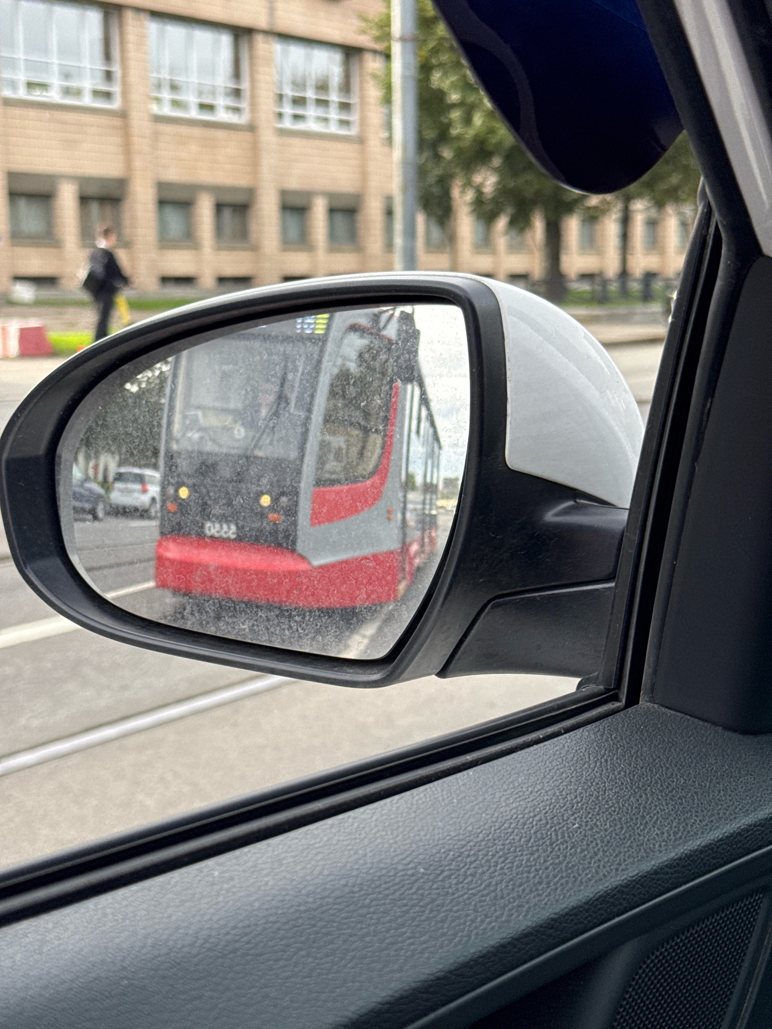 The image depicts a view from the interior of a car, looking towards the side-view mirror. The mirror reflects a tram with a red and gray exterior, its front lights illuminated. The tram appears to be on tracks, suggesting an urban environment. In the background, visible through the car window and beyond the mirror's reflection, is a light-colored building with multiple windows, and trees. A person in a dark suit is walking away from the camera on the sidewalk in front of the building. The car's interior, including the door panel and part of the car's roof, is also visible. The overall scene suggests a cloudy day in a city. The presence of trams and architectural style of the building are not specific enough to pinpoint a location in Saint Petersburg without further information.