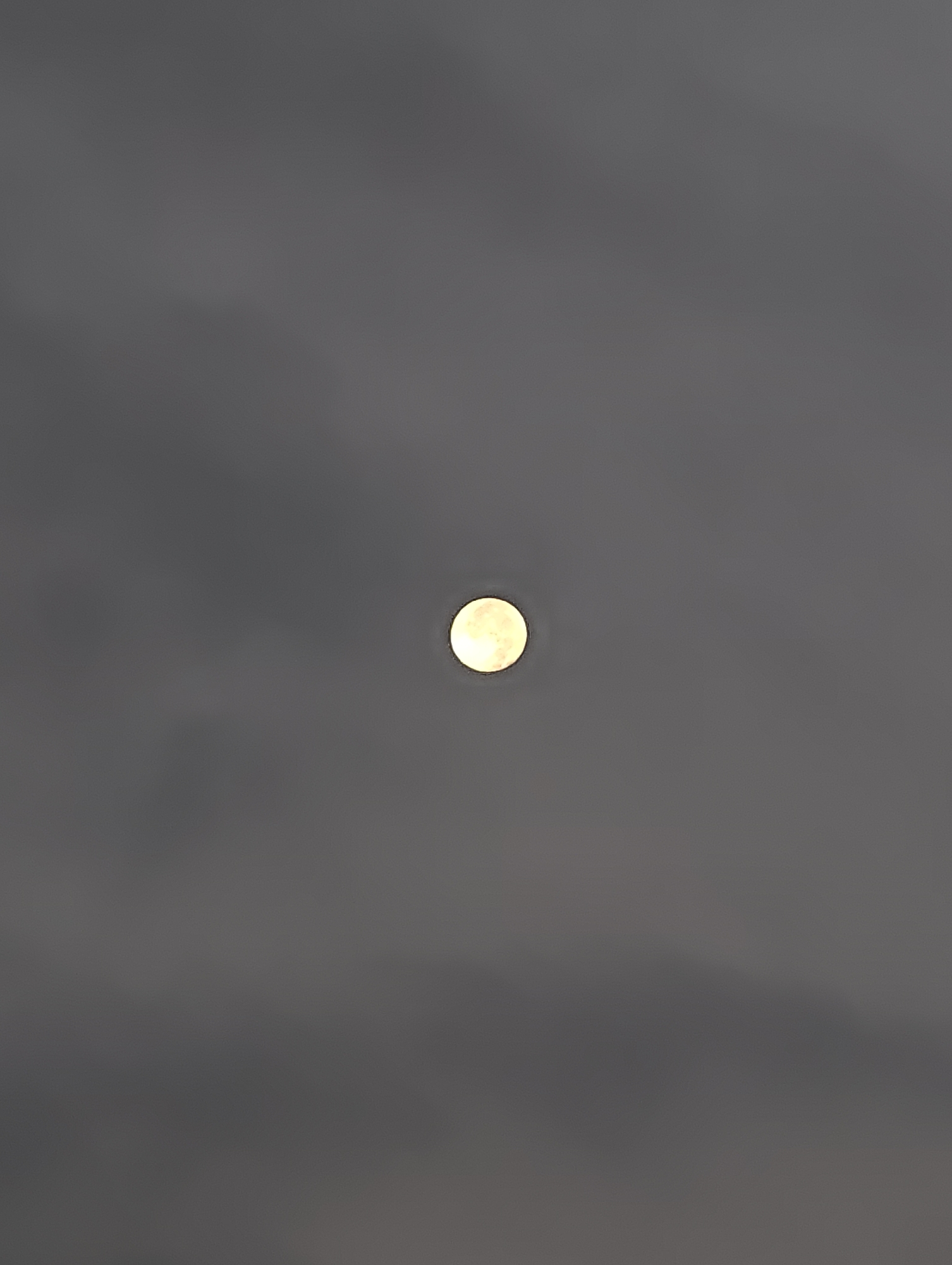 The image shows a full moon against a hazy, grey sky. The moon is a bright, pale yellow orb, slightly diffused by thin clouds or atmospheric haze. The overall color palette is muted, dominated by shades of grey and the pale yellow of the moon. There are no discernible landmarks or urban features visible in the image, and therefore, its specific location in Petah Tikva, Israel cannot be determined from the provided visual information. The scene is devoid of any people or specific objects, presenting a natural, celestial phenomenon.