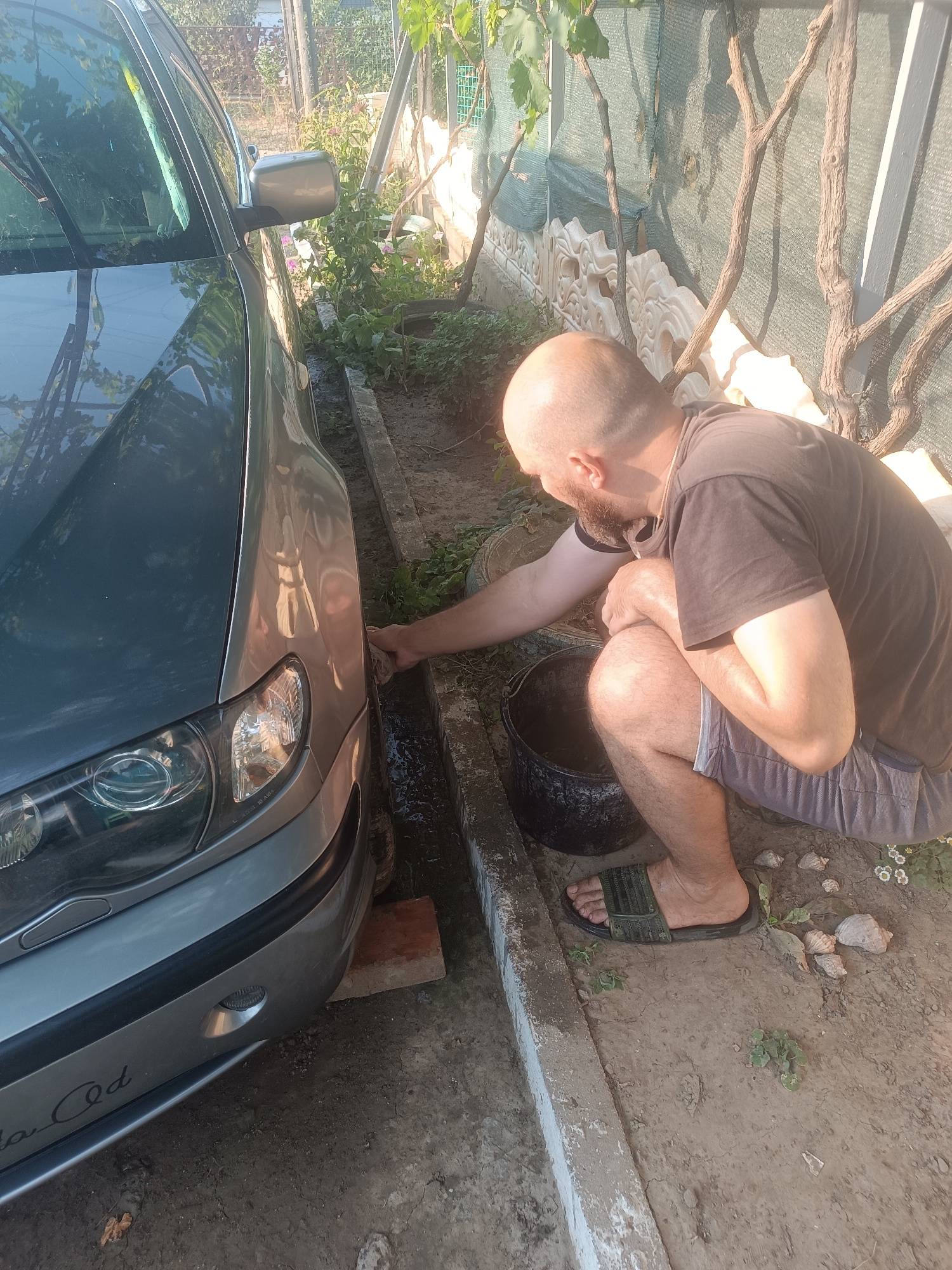 A bald, bearded man wearing a brown t-shirt and grey shorts is squatting next to a gray car. He is using a cloth to clean the side of the car, which is parked in a narrow dirt area bordered by a concrete channel and a fence. A black bucket is to his left. The scene appears to be in Vypasne, Ukraine, with vegetation and a fence visible in the background.