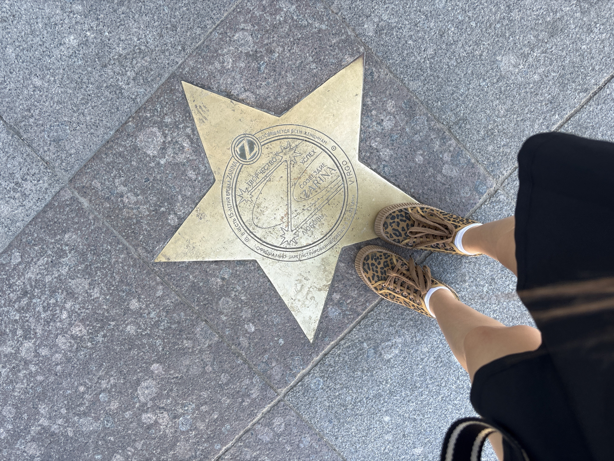 The image shows a close-up, high-angle view of a person's legs, wearing leopard-print sneakers and a black skirt, standing on a pavement. The person is positioned over a golden star embedded in the granite pavement. The star has inscriptions in Russian and English, indicating it is a dedication to the brand ZARINA on its 15th anniversary. The star also features astrological symbols and text related to "Virgo," "Love," "Creativity," "Success," and "Constellation ZARINA." The pavement itself is made of large granite slabs with visible seams. The lighting suggests it is daytime. This is likely located on Nevsky Prospekt in Saint Petersburg, Russia, where ZARINA has its Alley of Stars.